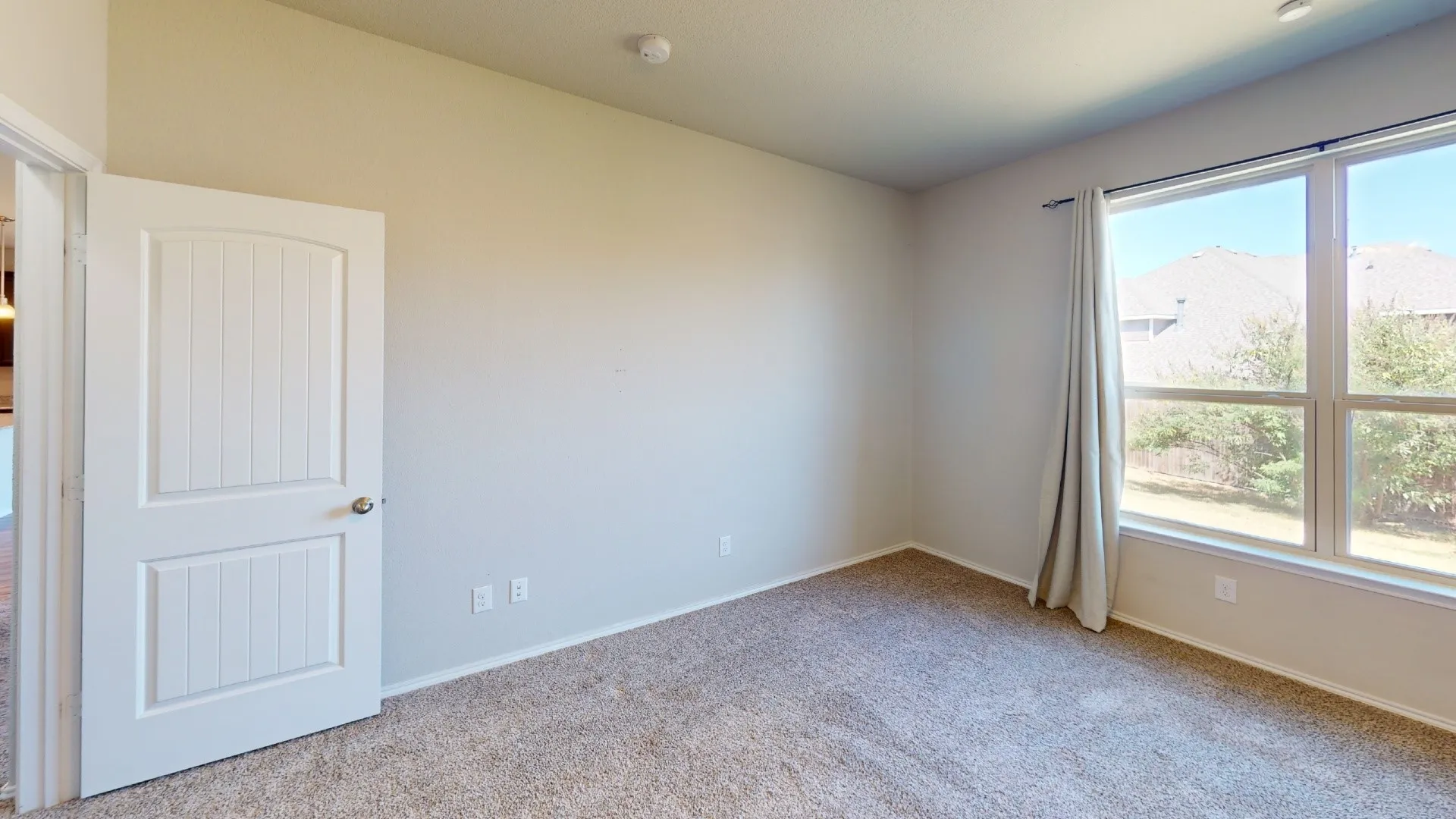 Carpeted room featuring a white paneled door, light-colored walls, and a large window overlooking outdoor greenery