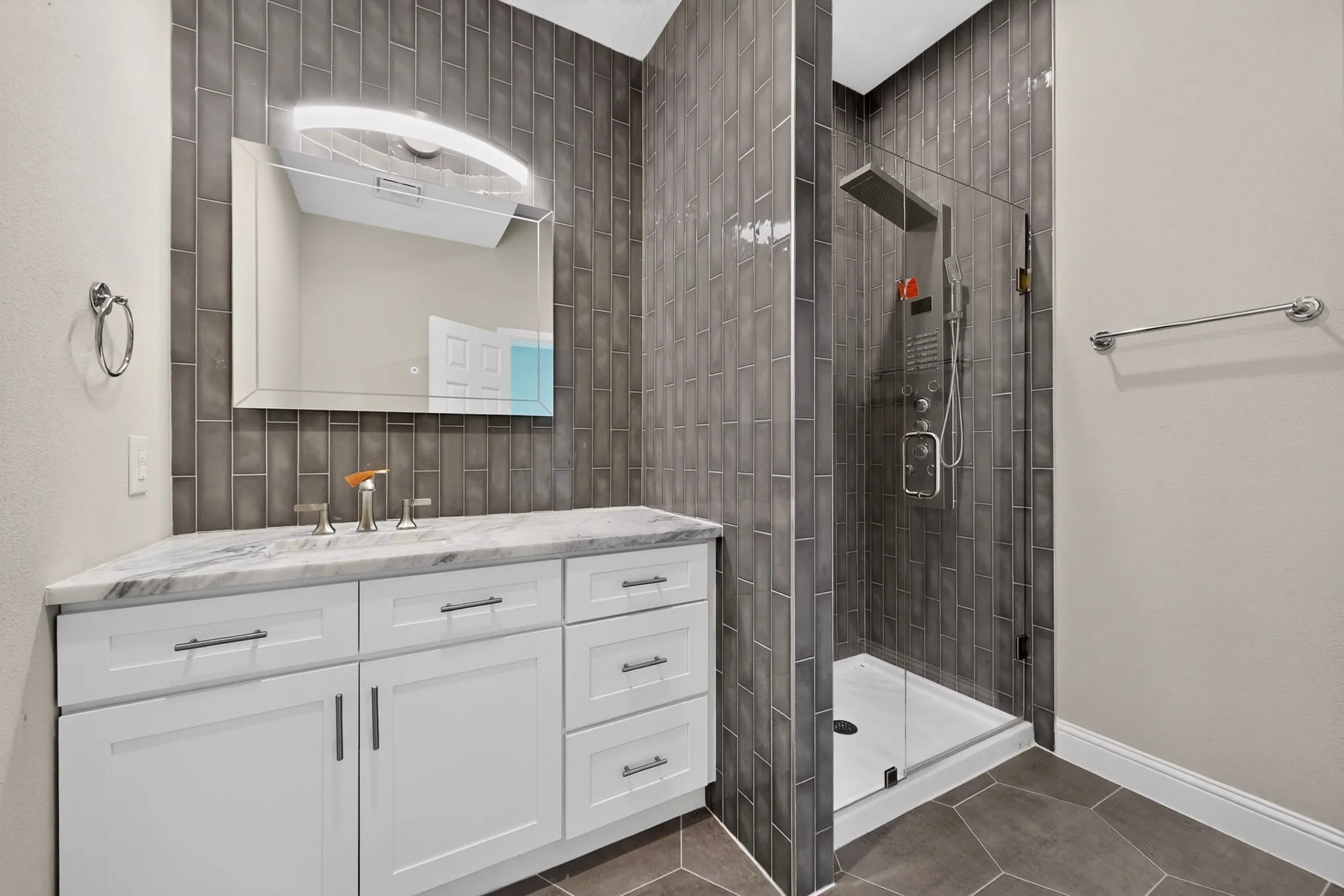 White shaker-style vanity with a light-colored countertop, wall-mounted mirror with integrated lighting, tiled shower with glass enclosure, and a multi-function shower system