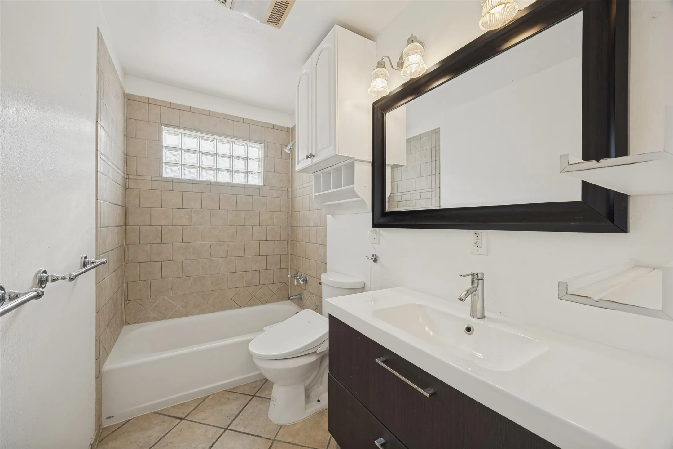 Bathroom featuring a dark wood-finish vanity with an integrated sink, a framed mirror, and a tiled shower/tub combination with a glass block window