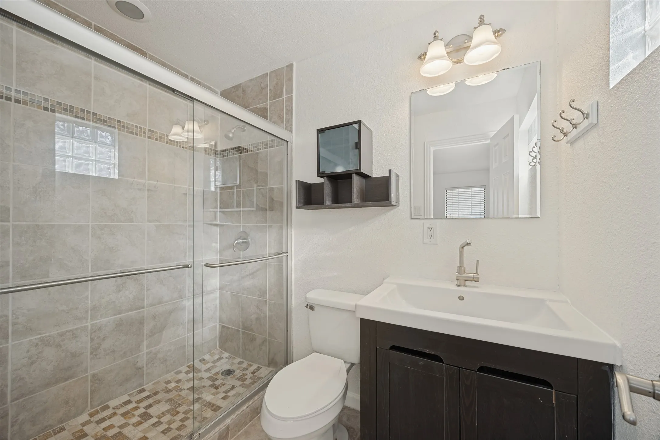 Glass-enclosed shower with tile surround and mosaic floor, dark wood-finish vanity with an integrated sink, and a rectangular mirror with vanity lighting