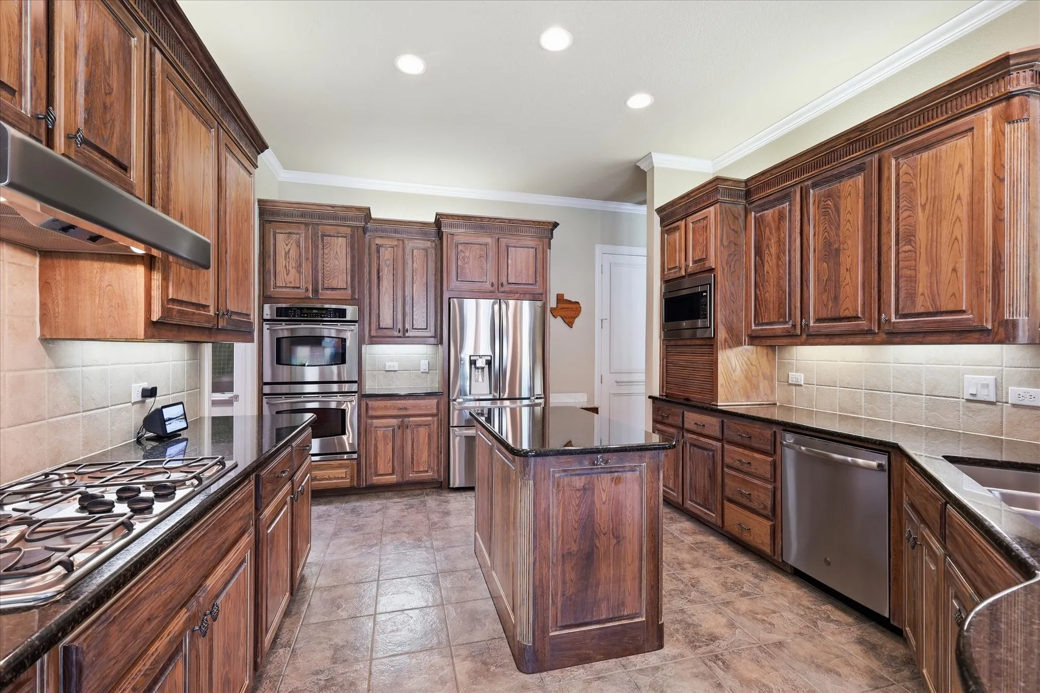 Wood-finish cabinetry, dark countertops, a central island, integrated stainless steel appliances, and tile flooring