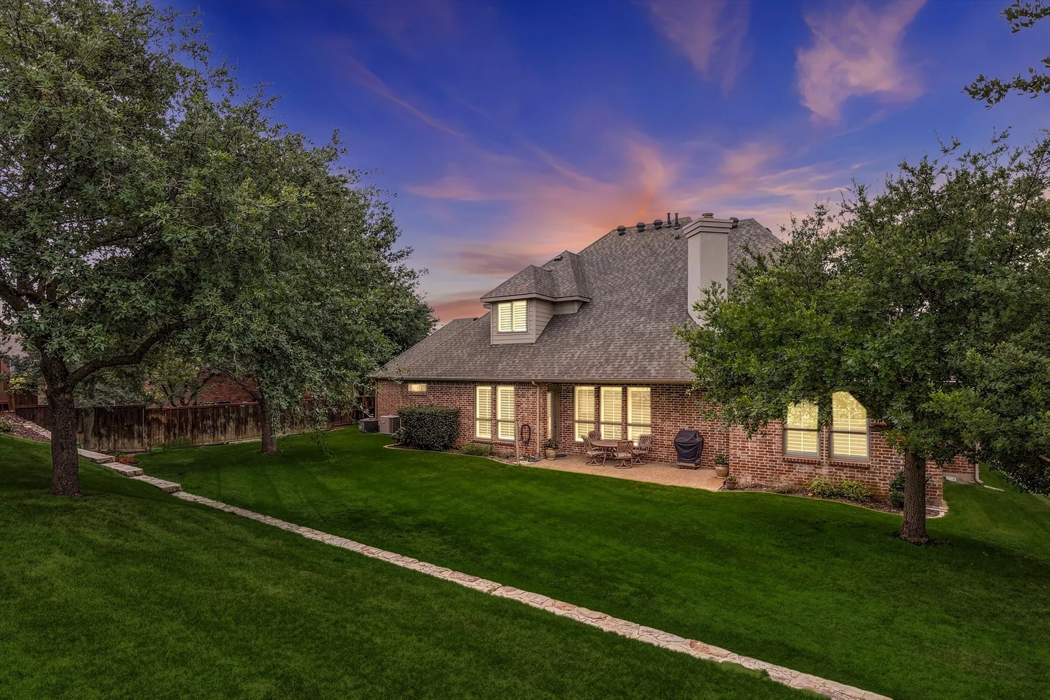 Brick exterior residence with a shingled roof, a ground-level patio, multiple windows, and a stone-edged lawn path