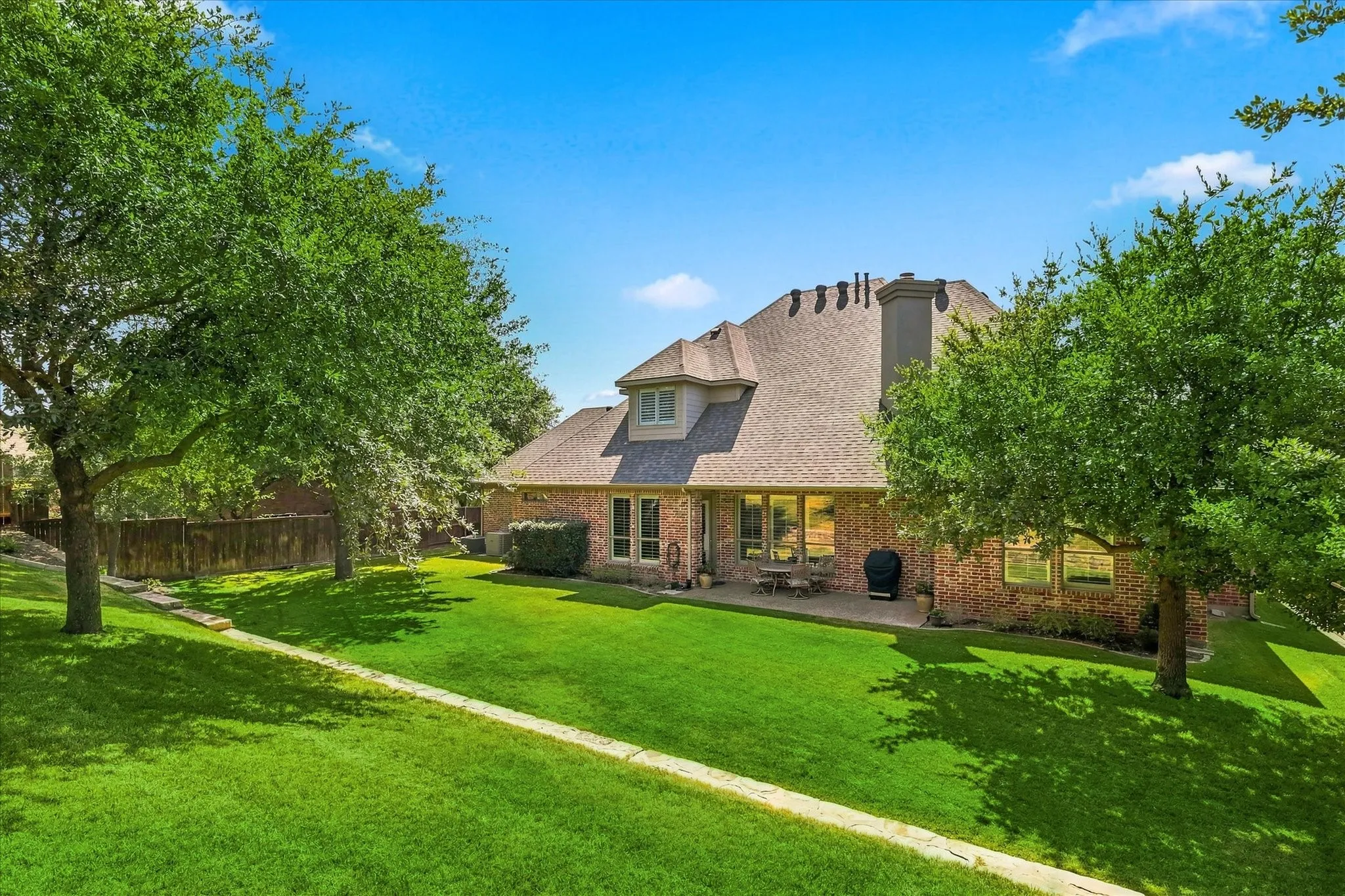 Expansive backyard featuring a brick exterior, a manicured lawn, a concrete patio, and mature trees