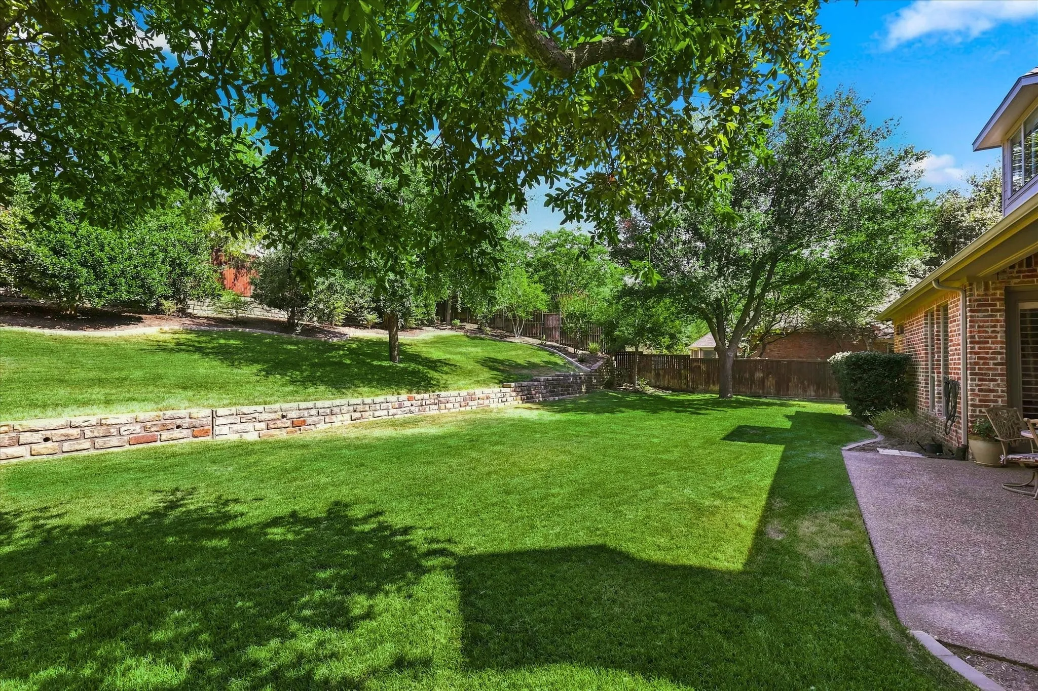 Expansive tiered backyard featuring a manicured lawn, established trees, and a stone retaining wall