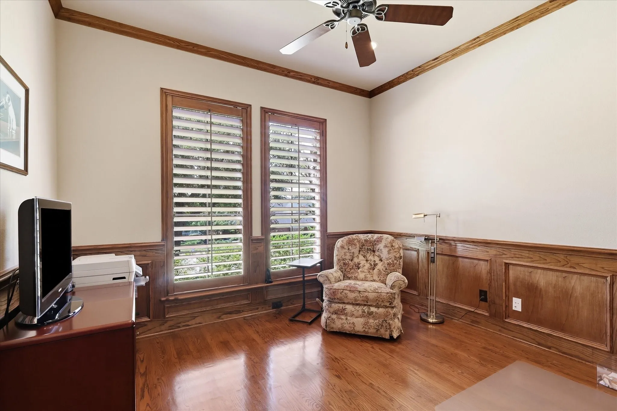 Hardwood flooring, wood paneling, wood window frames with plantation shutters, crown molding, and a ceiling fan with integrated lighting