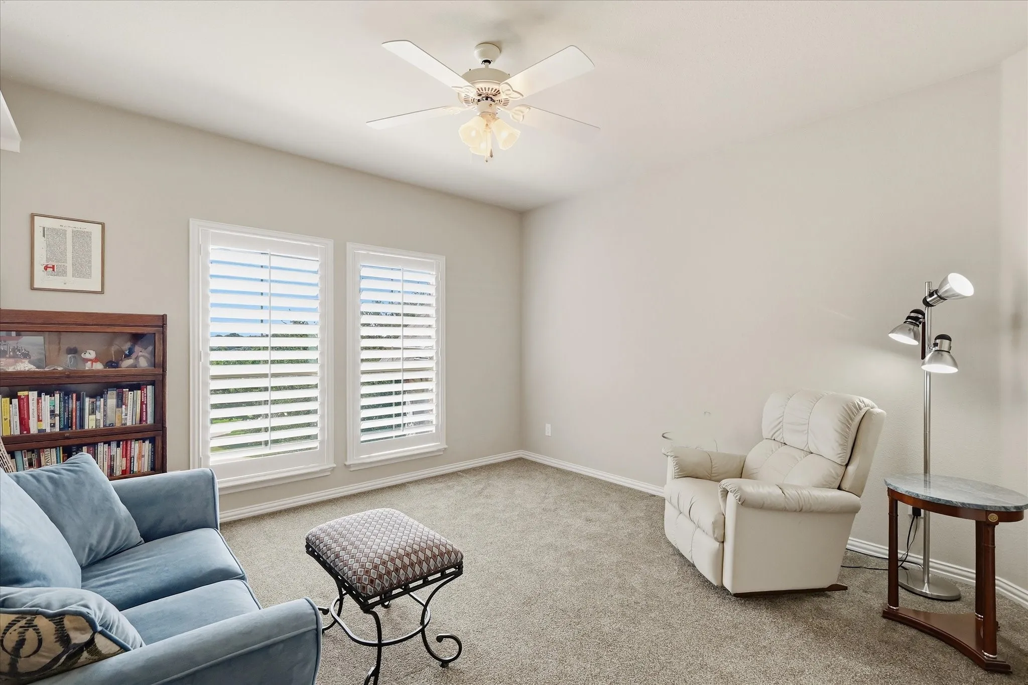 Neutral wall paint, ceiling fan with integrated lighting, two windows with white plantation shutters, and light-colored carpeting