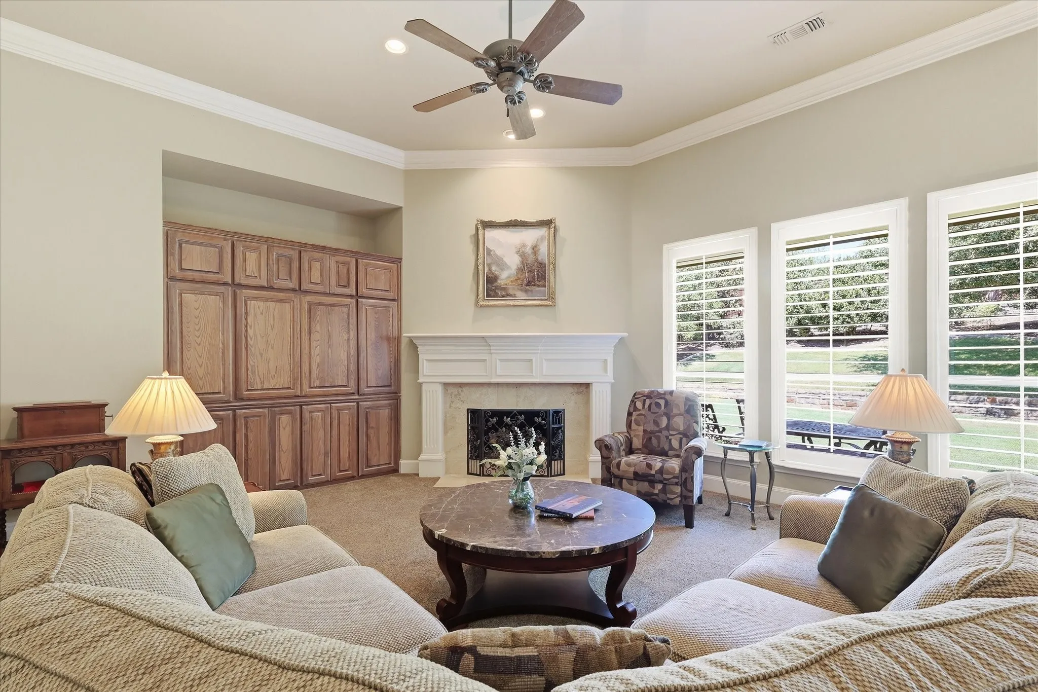 Recessed wood cabinetry, white fireplace with hearth, three large windows with plantation shutters, ceiling fan, and crown molding