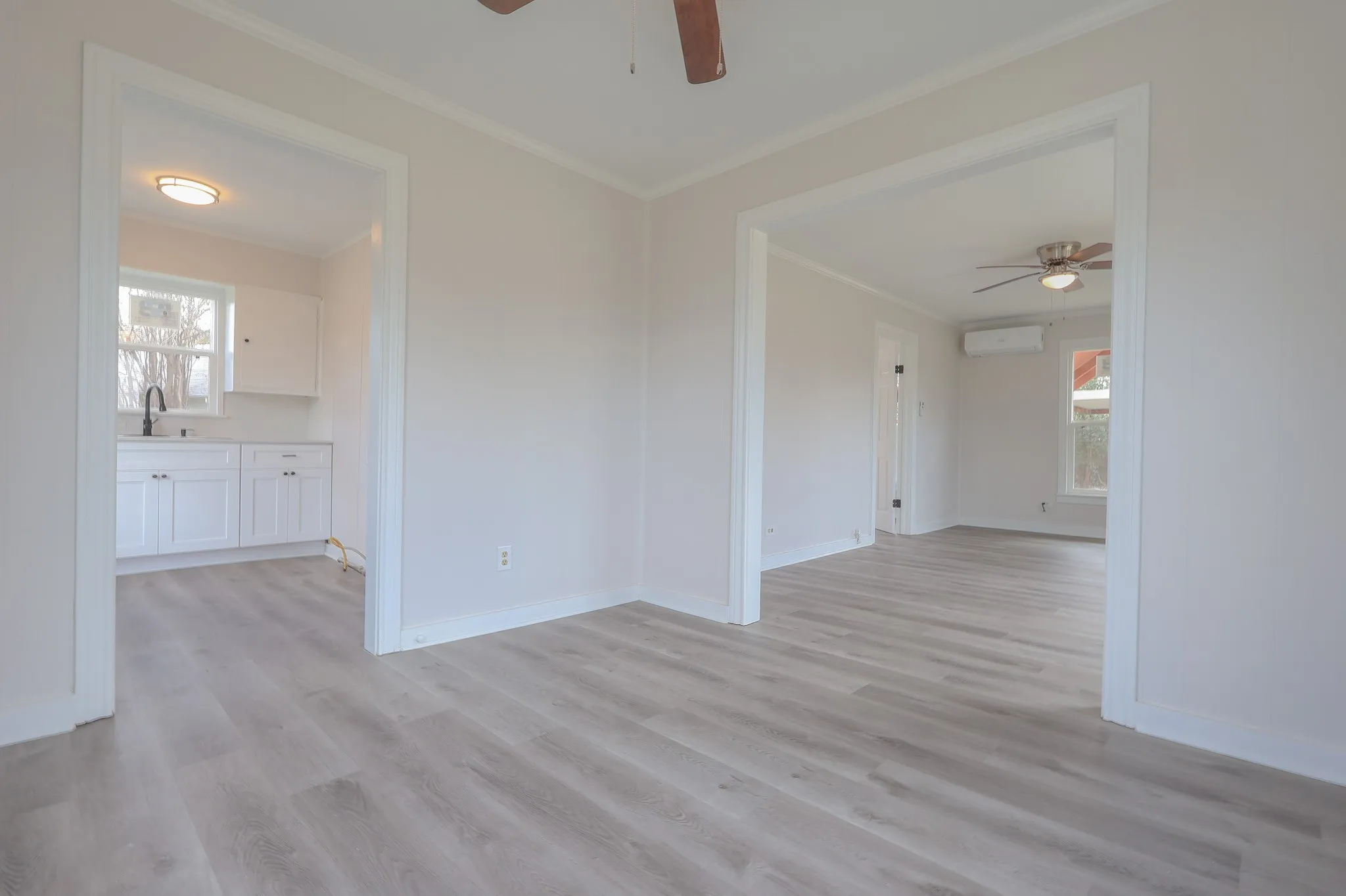 Light wood-finish flooring throughout, white walls with crown molding, white kitchen cabinetry with a dark faucet, and a ceiling fan