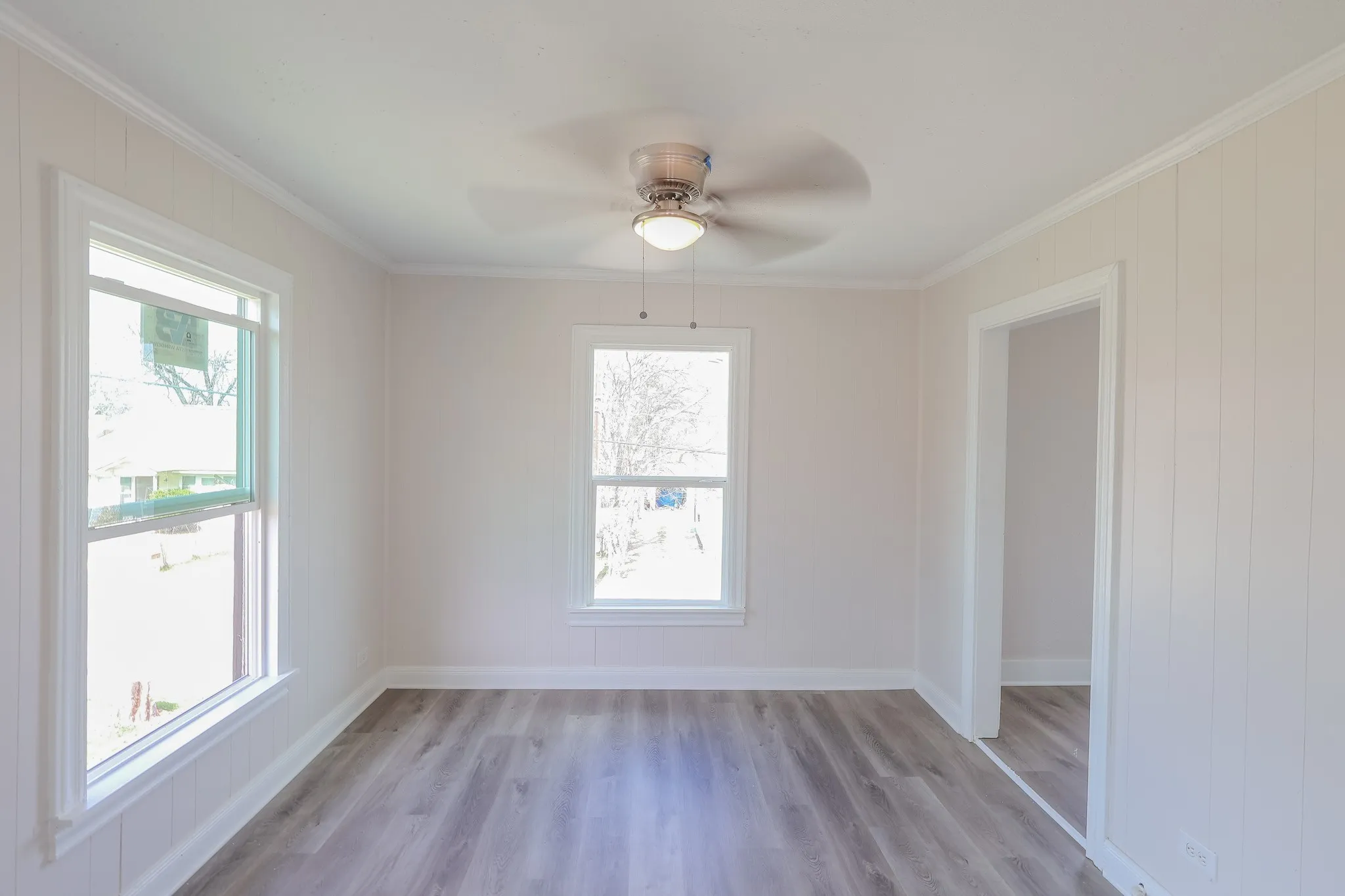 Wood-finish flooring, two large windows, white wall paneling, crown molding, and a ceiling fan with integrated lighting