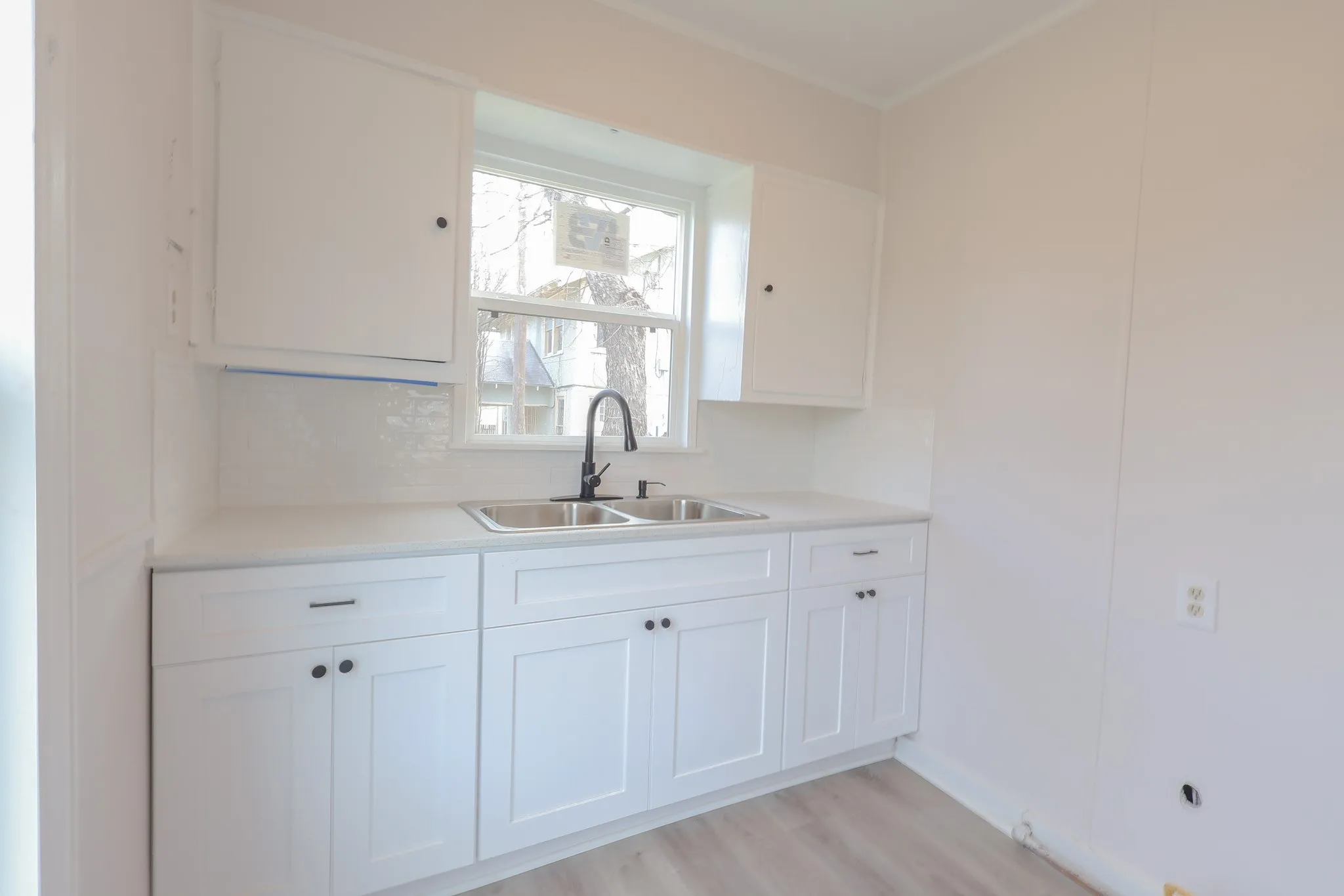 White shaker-style cabinetry with dark hardware, dual stainless steel sinks with a gooseneck faucet, light-toned countertops, white subway tile backsplash, and wood-finish flooring
