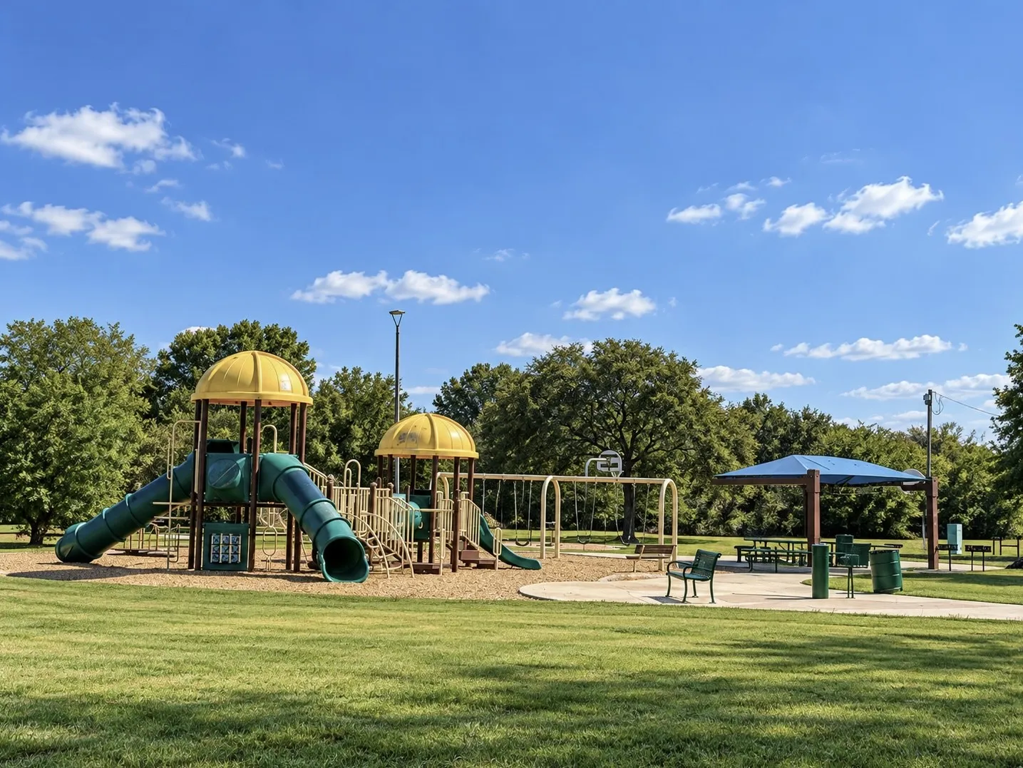 Community playground with multiple slides, swings, and sheltered picnic area, surrounded by green space and mature trees
