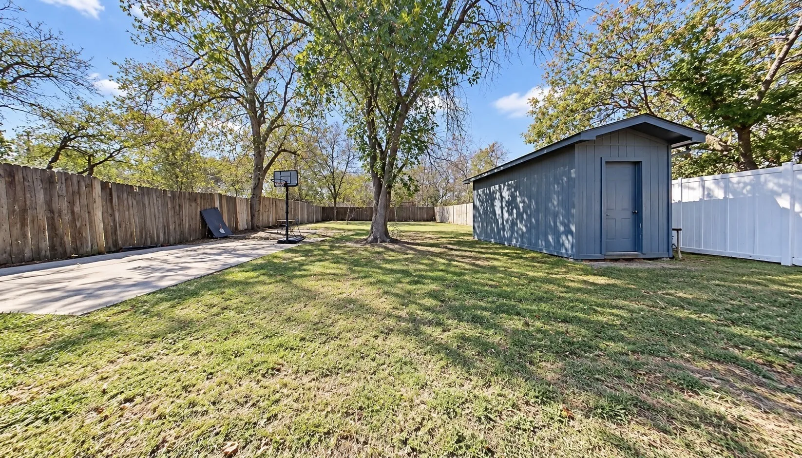 Expansive yard featuring a large concrete pad, mature trees, wood privacy fencing, and a storage shed with a gabled roof
