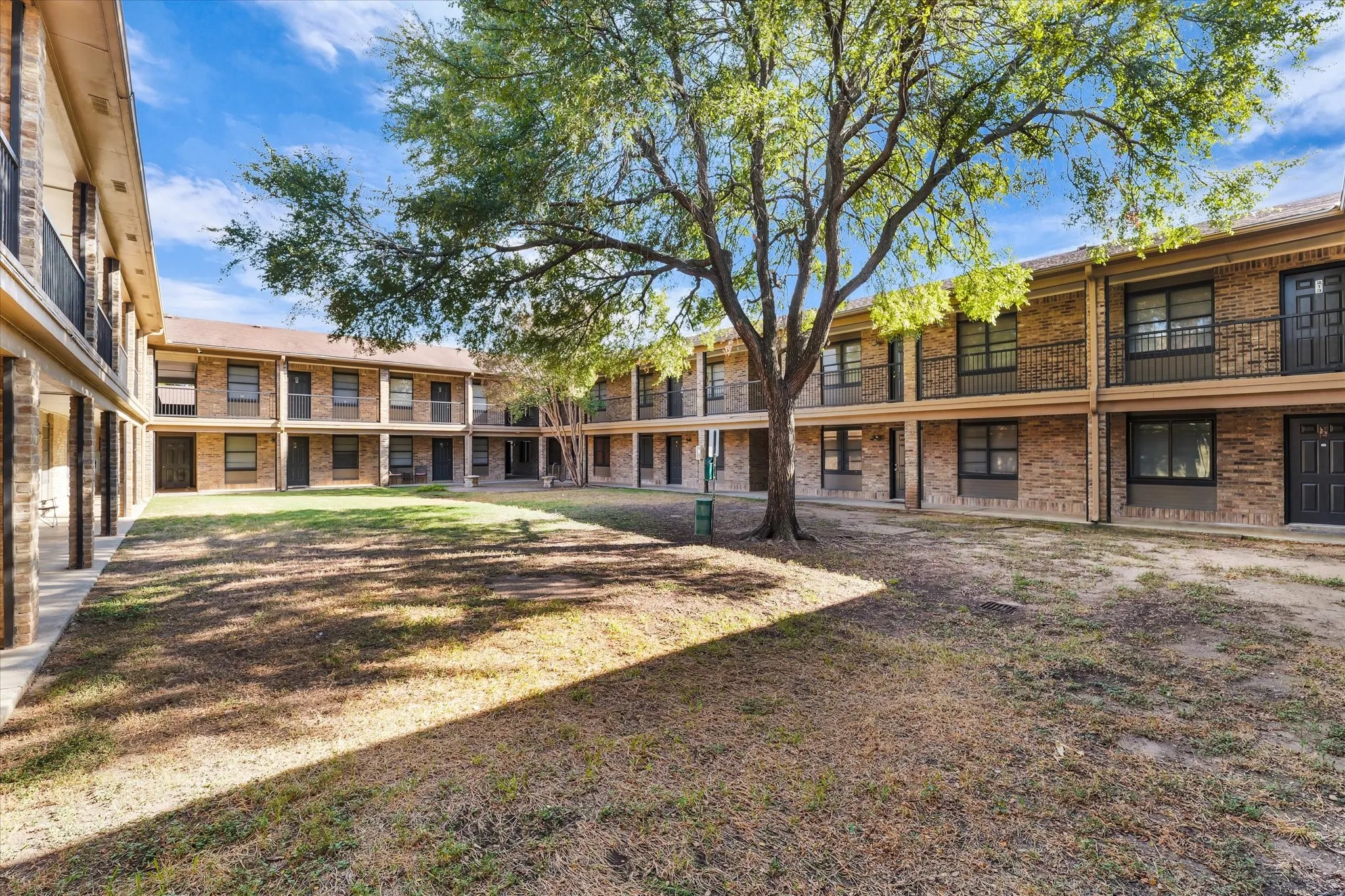 Central courtyard featuring a mature tree, surrounding two-story buildings with brick exteriors, and upper-level balconies with wrought iron railings