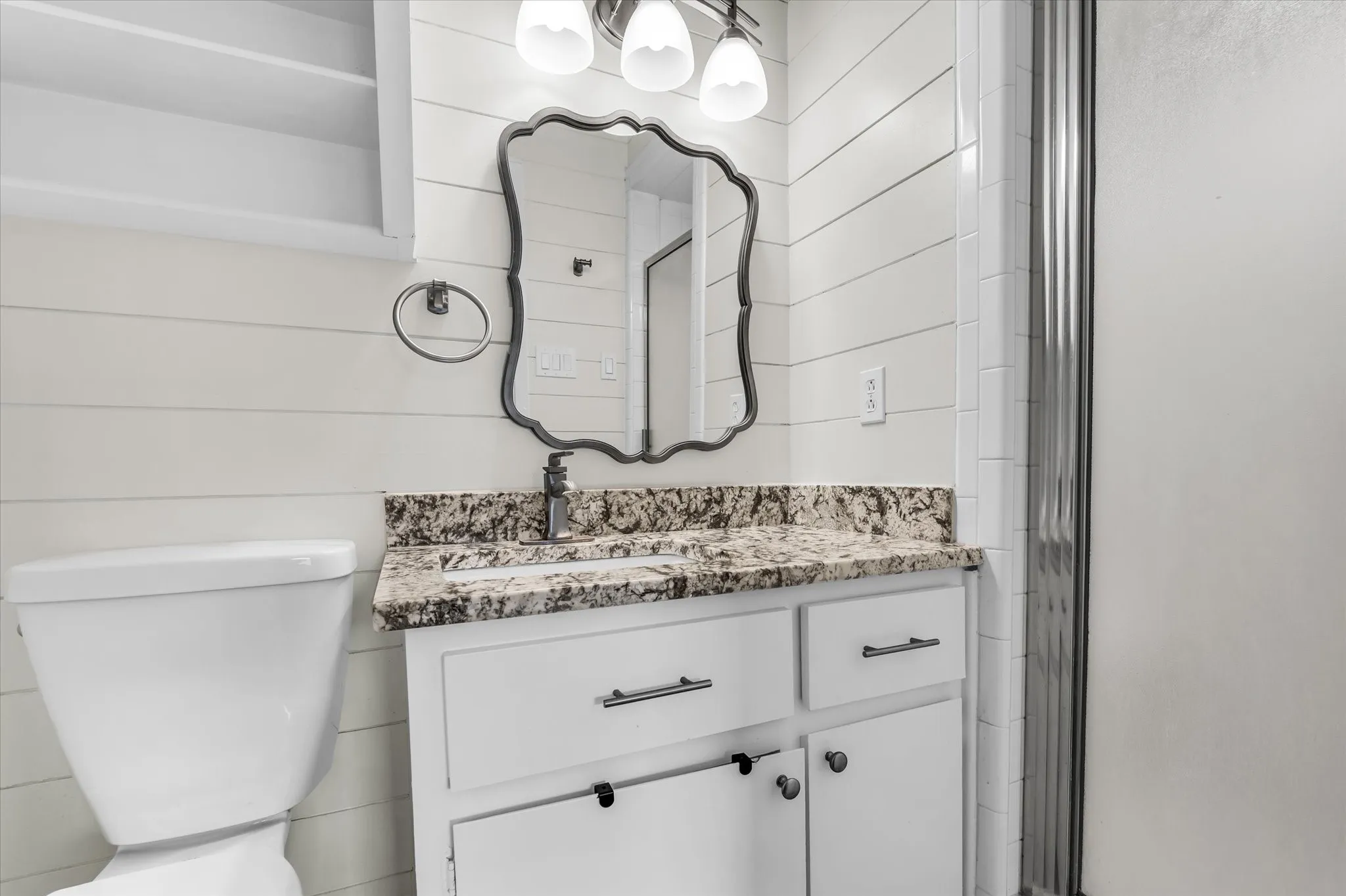 White shiplap walls, vanity with granite countertop, white cabinetry with dark hardware, framed mirror, and a modern light fixture
