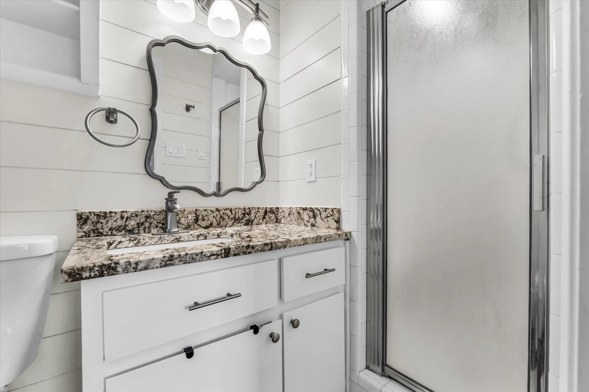 White vanity with a stone countertop and integrated sink, framed mirror, wall-mounted vanity lighting, and a glass-enclosed shower