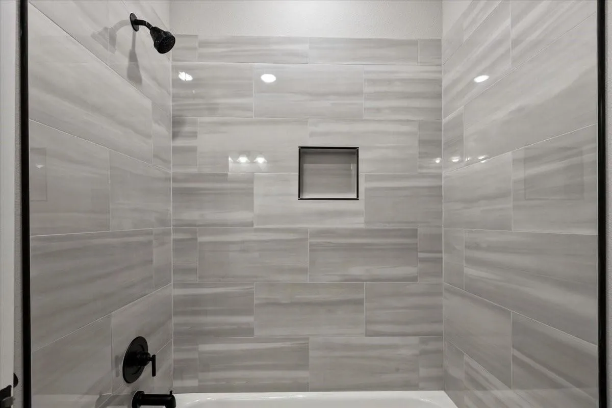 Bathtub with matte black fixtures, recessed shower niche, and light-toned horizontal tile surround