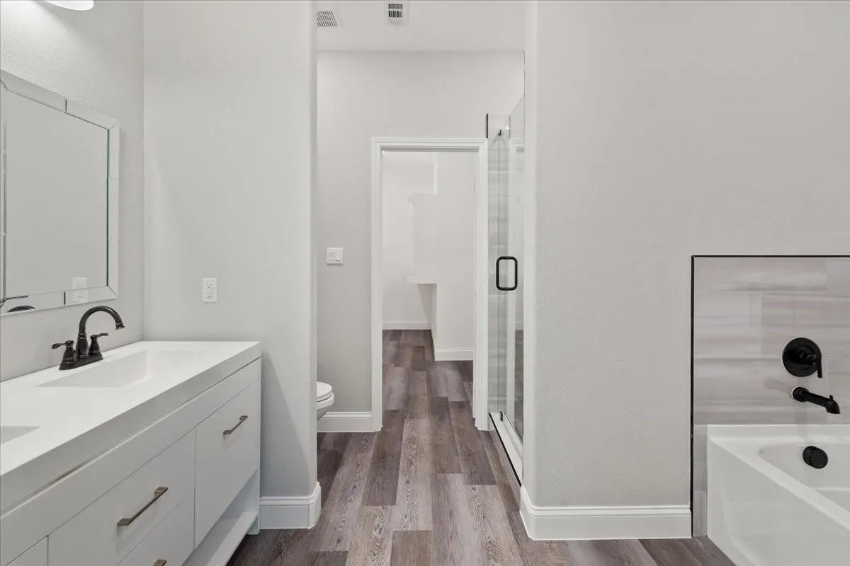 White vanity with matte black faucet, framed mirror, glass-enclosed shower, white bathtub with black fixtures, and wood-finish flooring