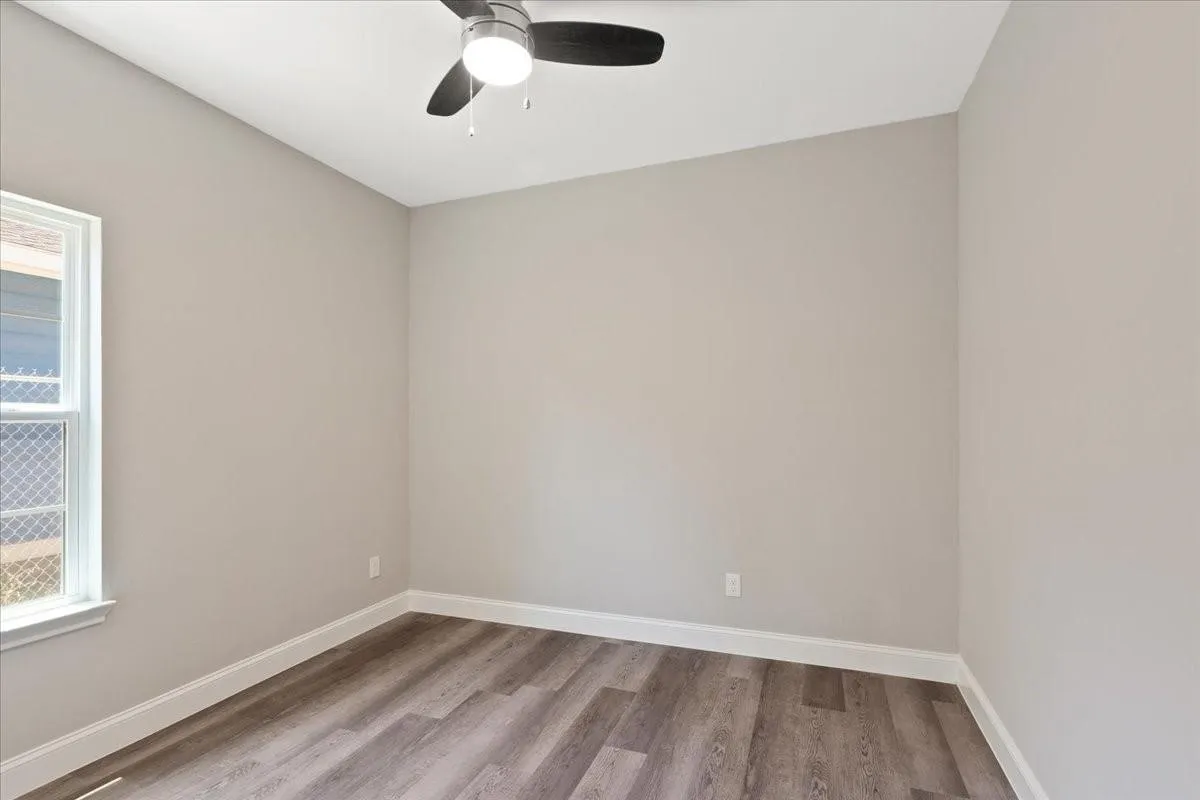 Light-filled room featuring wood-finish flooring, neutral wall paint, white trim, a single window, and a ceiling fan with integrated lighting