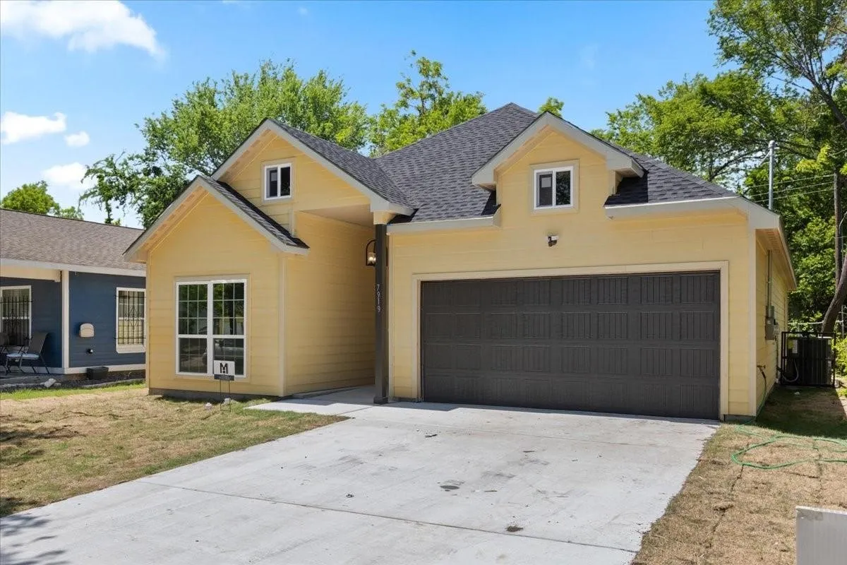 Yellow exterior with dark shingle roof, attached garage with dark panel door, dormer windows, and concrete driveway