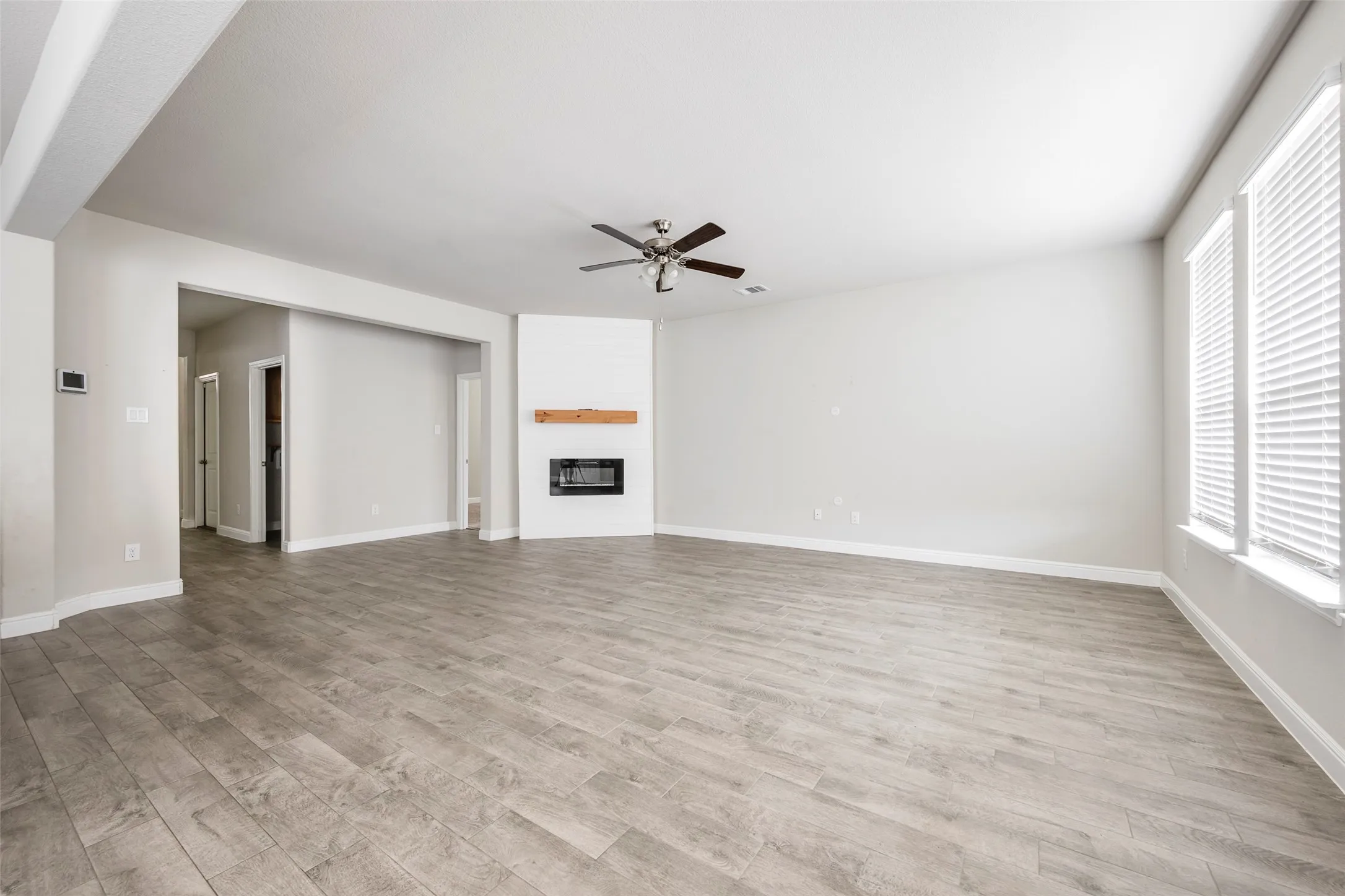 Spacious room featuring wood-finish flooring, a contemporary fireplace with a wood mantle, a ceiling fan, and large windows with blinds