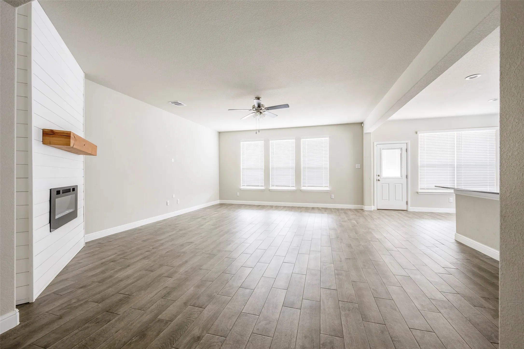 Spacious living area featuring wood-finish flooring, a fireplace with a wood mantle, multiple windows, a ceiling fan, and a light-colored entry door