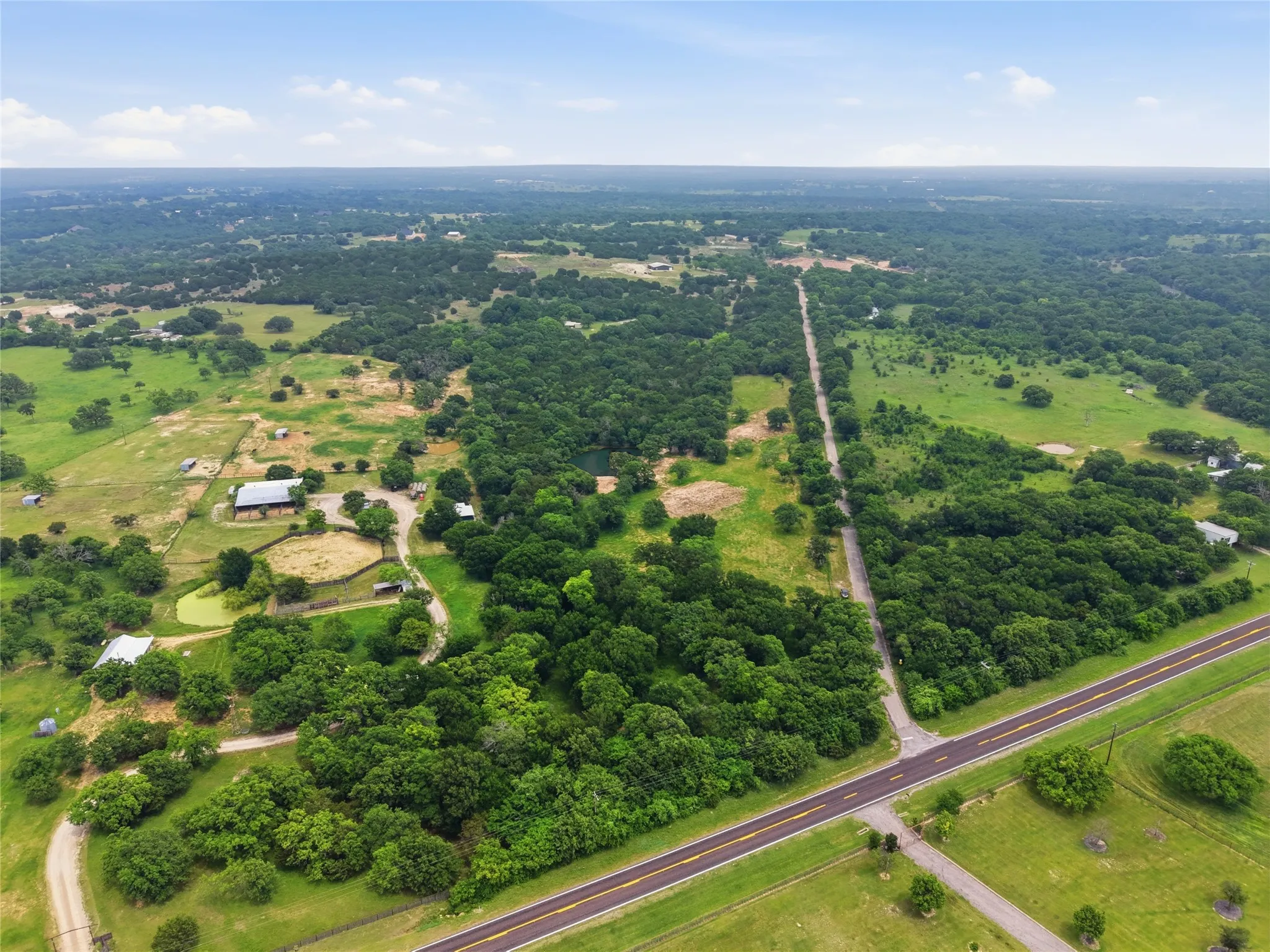 Expansive aerial landscape featuring a paved road, dense tree coverage, open green fields, a visible pond, and multiple structures