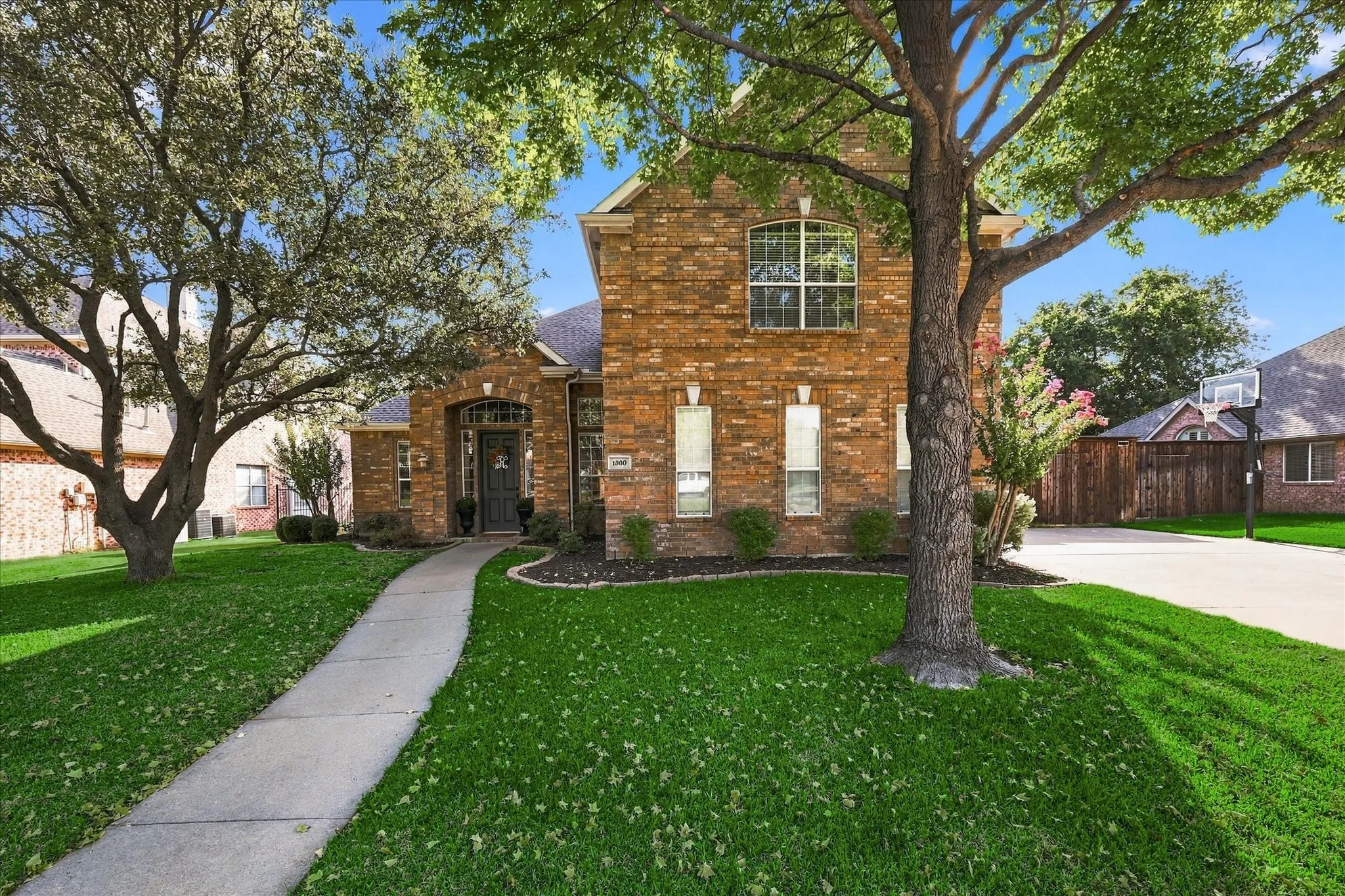 Brick exterior with an arched entry, mature trees, a well-maintained lawn, and a concrete driveway