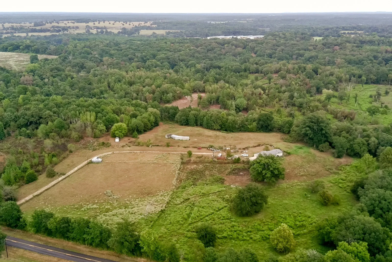 Expansive property featuring verdant tree coverage, open grassy areas, visible structures, and a partial roadway