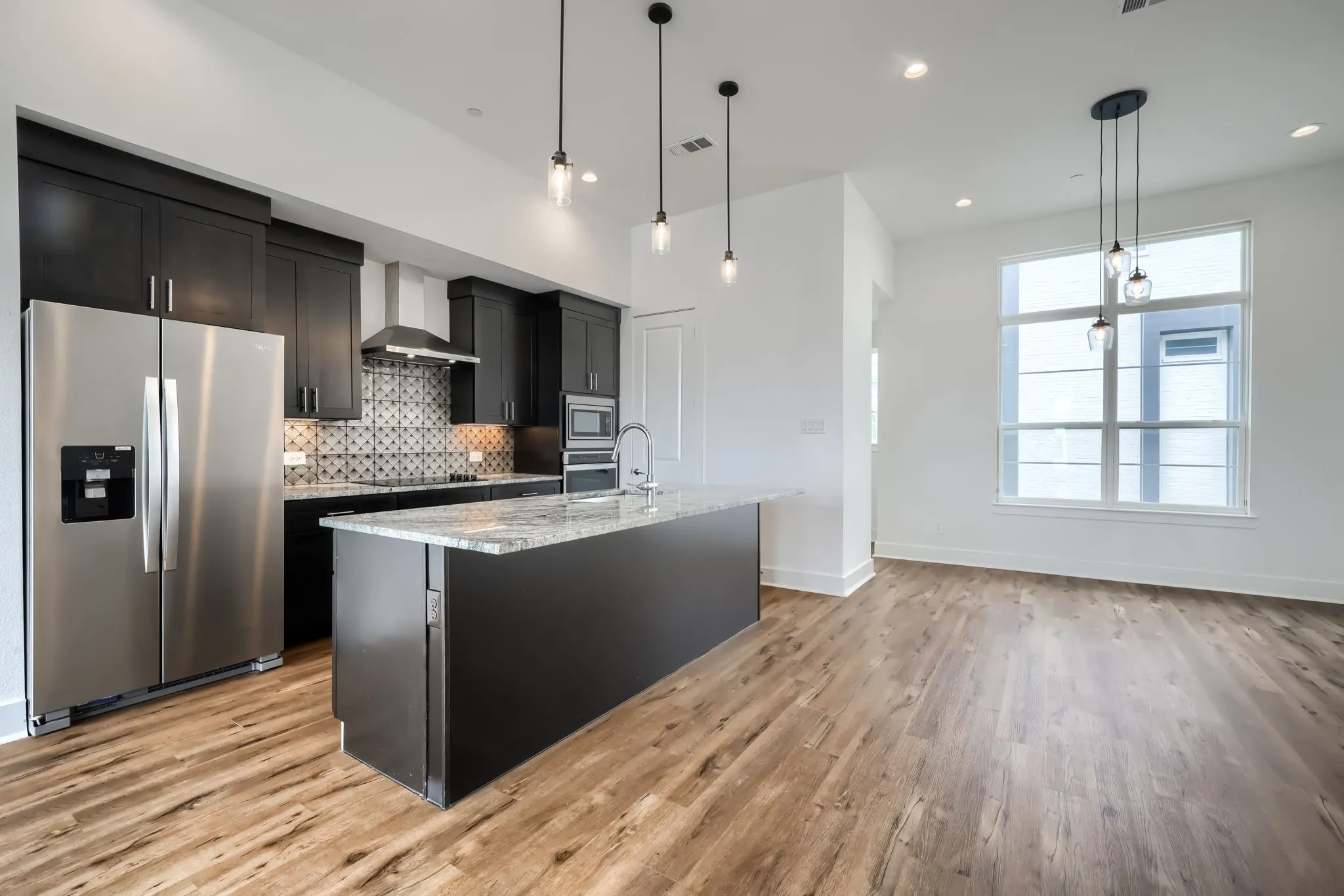 Open-concept layout featuring wood-finish flooring, a kitchen island with stone countertop, stainless steel appliances, dark cabinetry, and pendant lighting