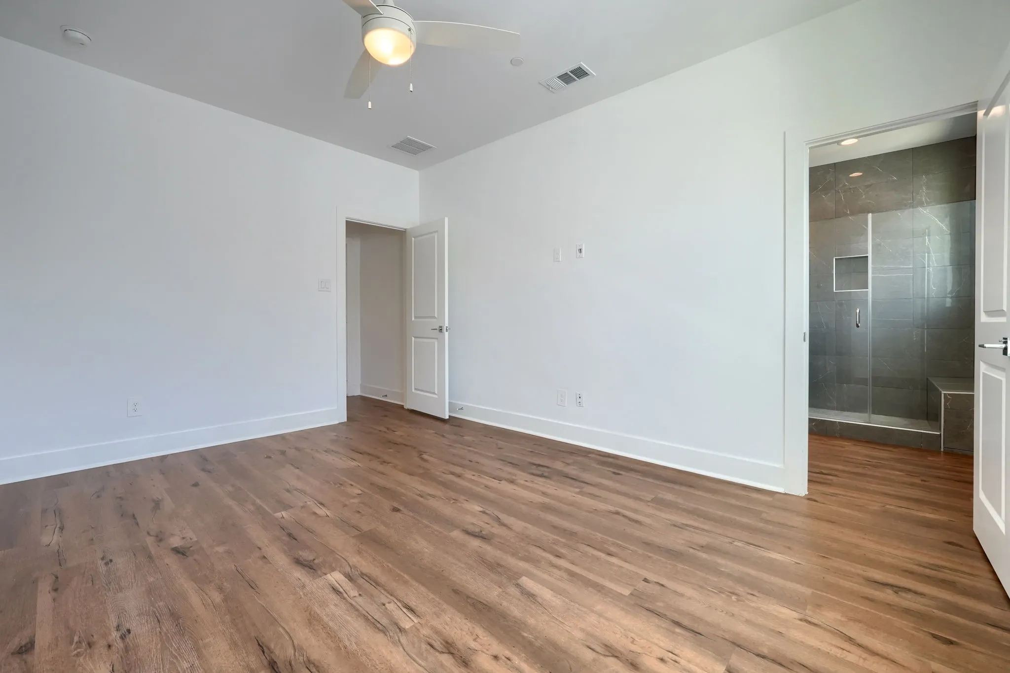 Wood-finish flooring throughout, white walls, ceiling fan with integrated lighting, and a walk-in shower with a glass enclosure and bench seating