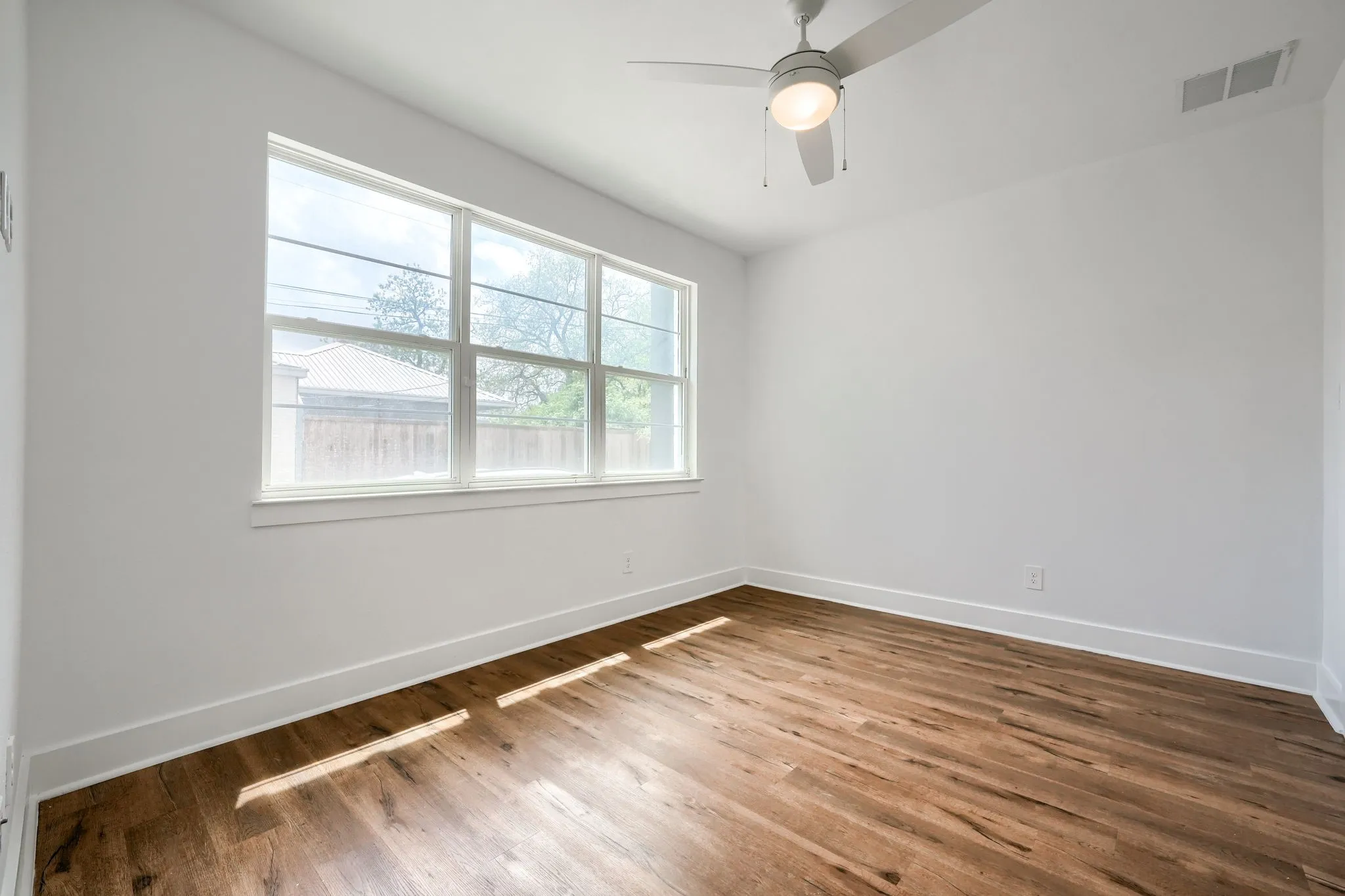 Wood-finish flooring with white walls, large multi-pane window, and a ceiling fan with integrated lighting