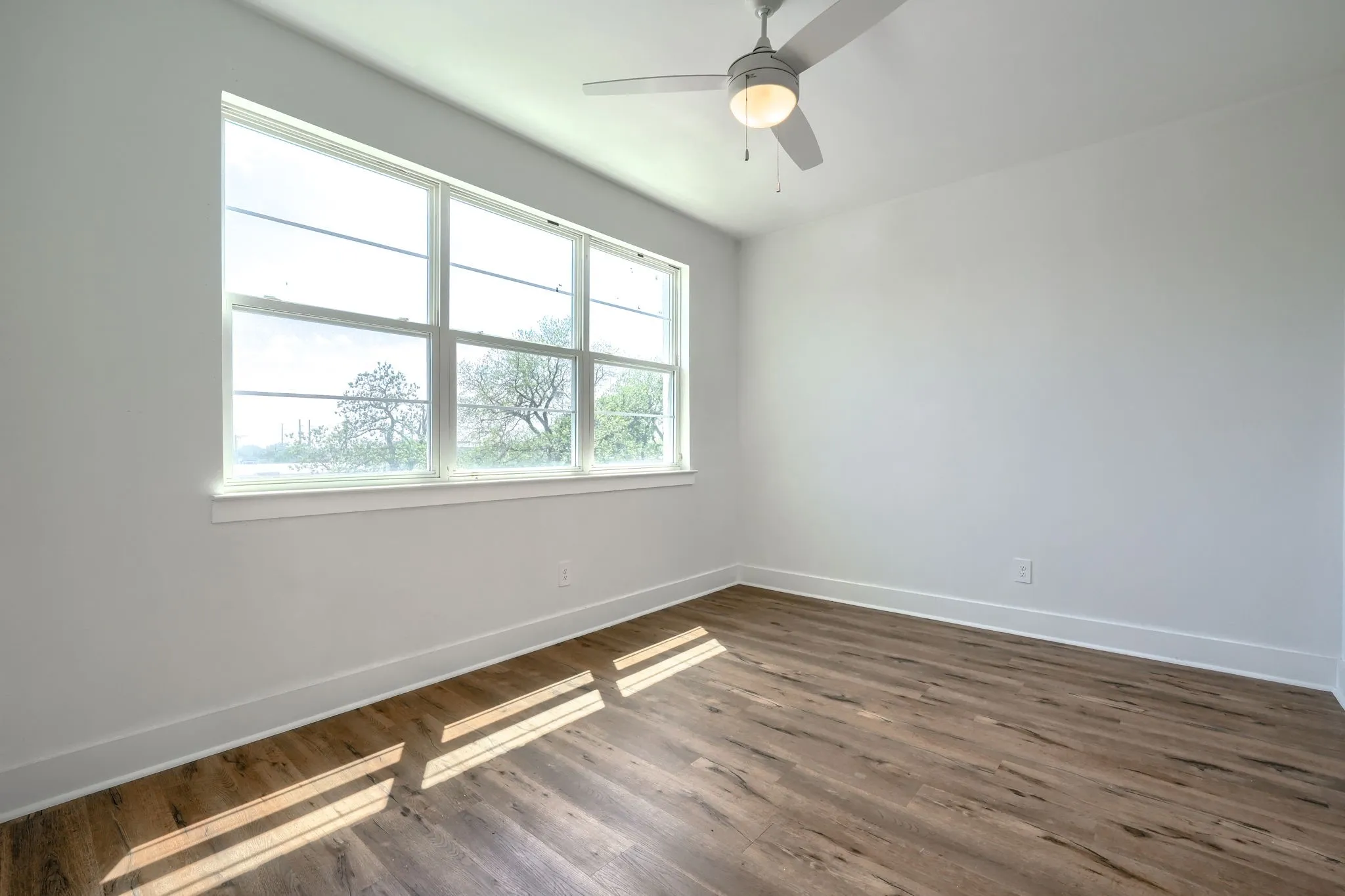 Wood-finish flooring, white walls, multiple windows, and a ceiling fan with integrated lighting