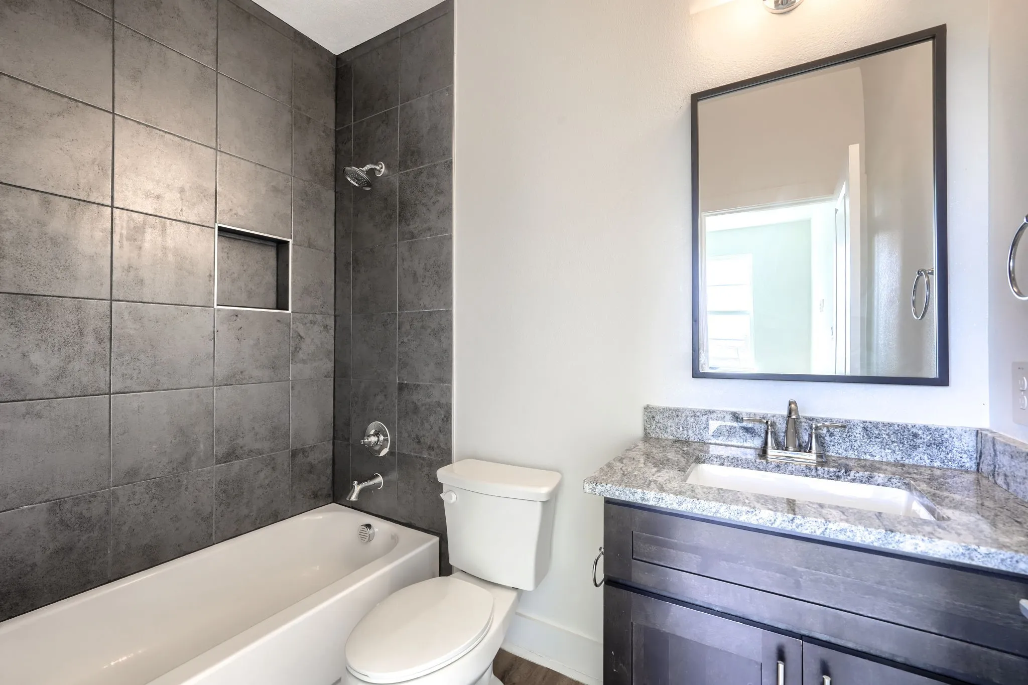 Full-height shower tiling with integrated niche, white bathtub, dark vanity cabinet with integrated sink, stone countertop, and framed mirror