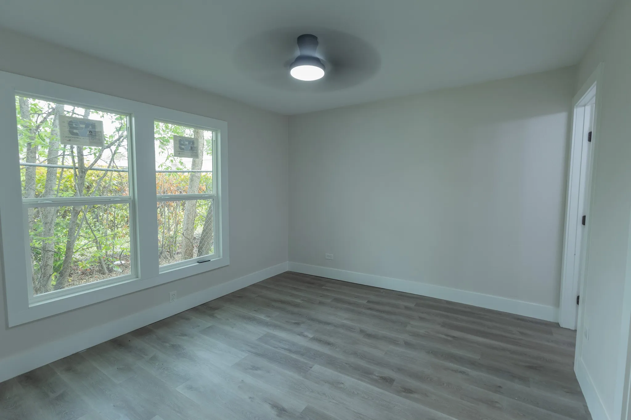 Neutral wall paint, wood-finish flooring, two large windows, white door frame, and a flush-mount ceiling light fixture