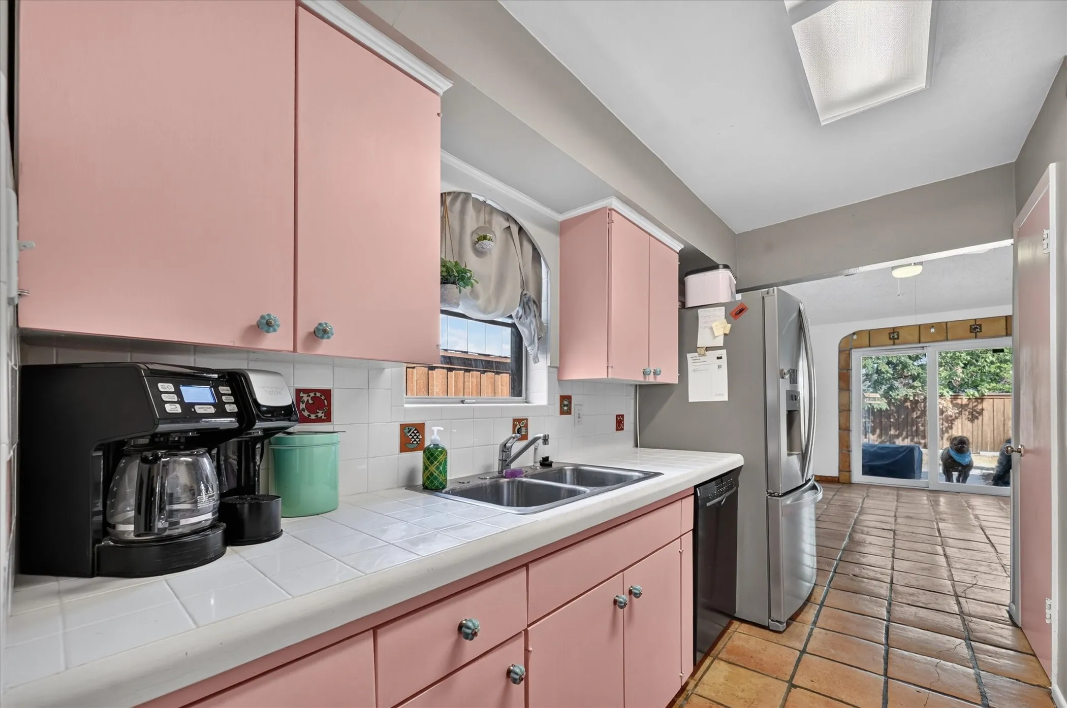 Pink cabinetry with decorative pulls, integrated dual basin sink, white tile countertops and backsplash, stainless steel refrigerator, and terracotta tile flooring