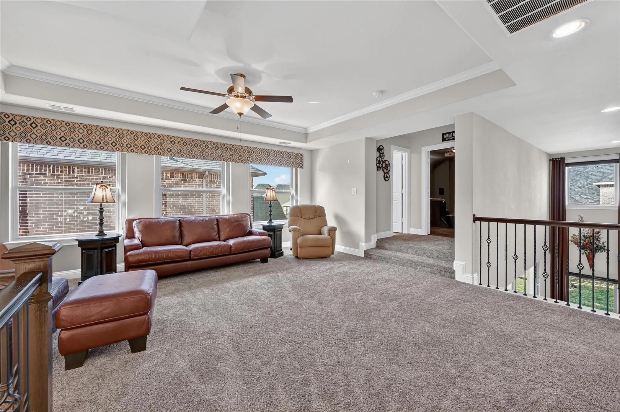 Spacious carpeted room featuring a ceiling fan, tray ceiling, multiple windows, and a black wrought iron railing
