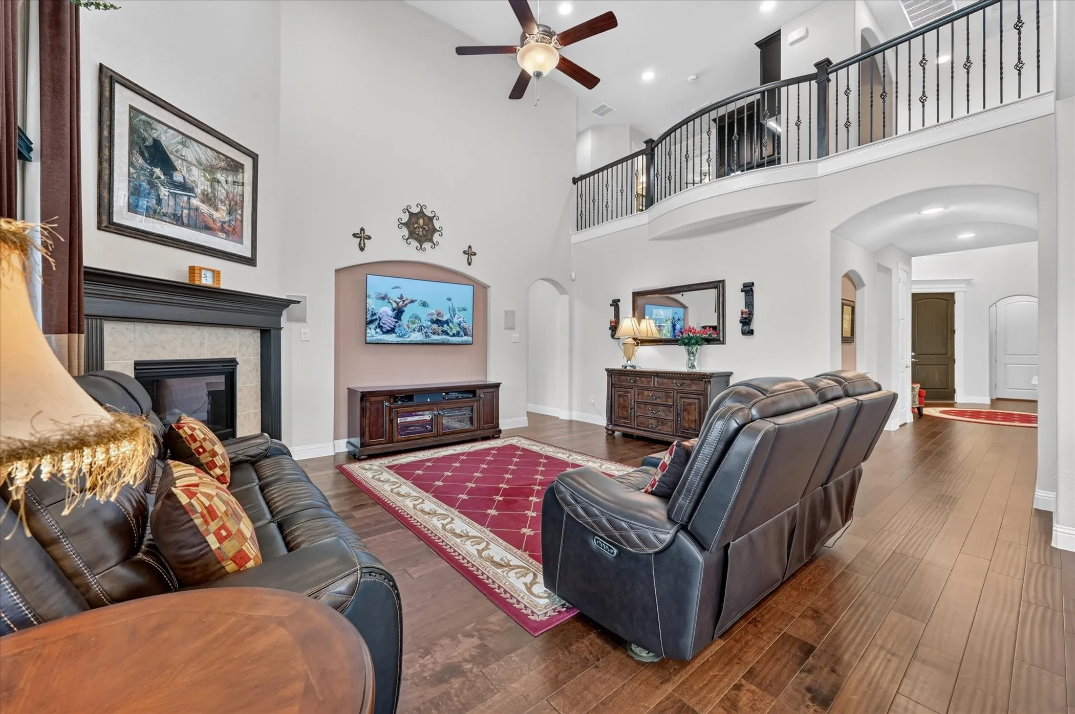 Two-story living area featuring wood-finish flooring, a fireplace with a mantel, a ceiling fan, and a second-story balcony with an iron railing