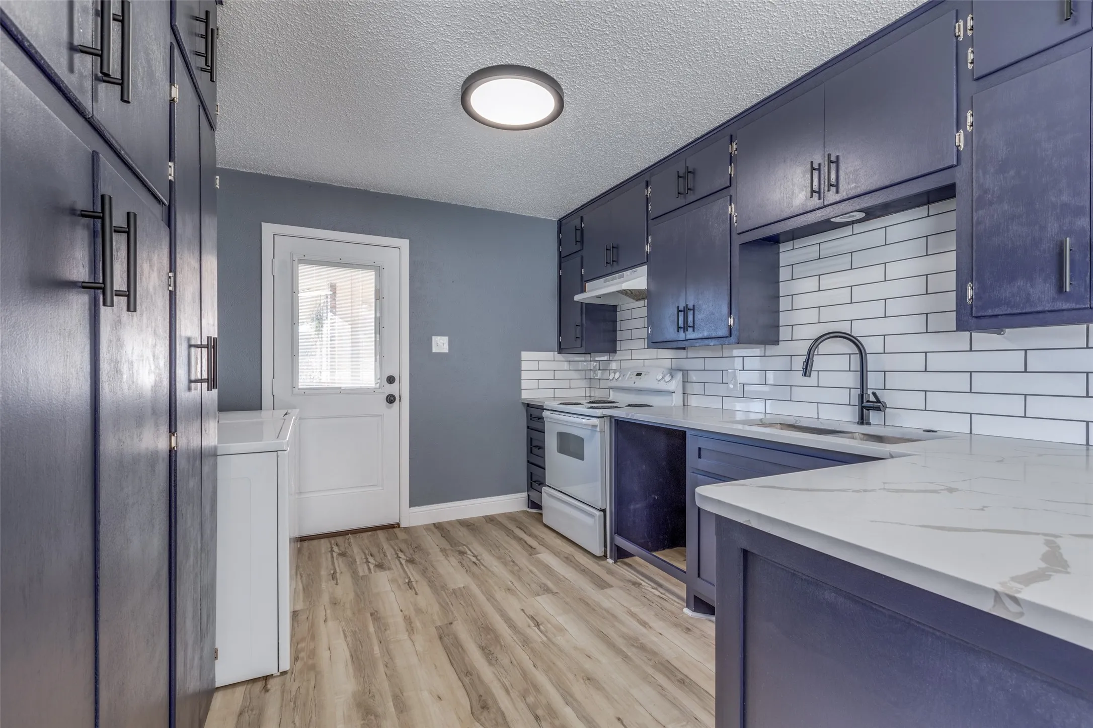 Kitchen featuring dark cabinetry with matte black hardware, wood-finish flooring, white subway tile backsplash, white countertops, and a full lite door
