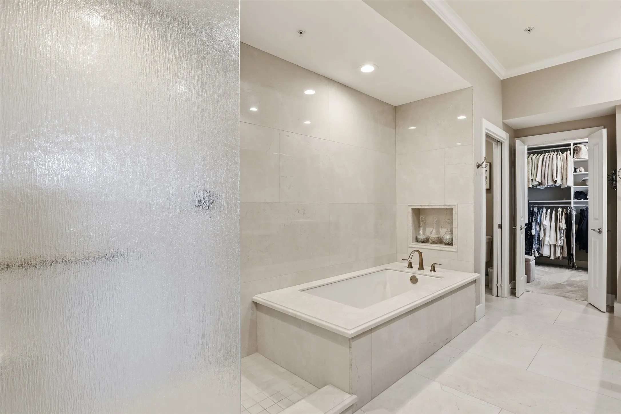 Spa-inspired primary bathroom featuring a freestanding soaking tub with integrated deck, frameless glass shower enclosure with textured wall panels, recessed ceiling lighting, and polished stone-finish flooring throughout
