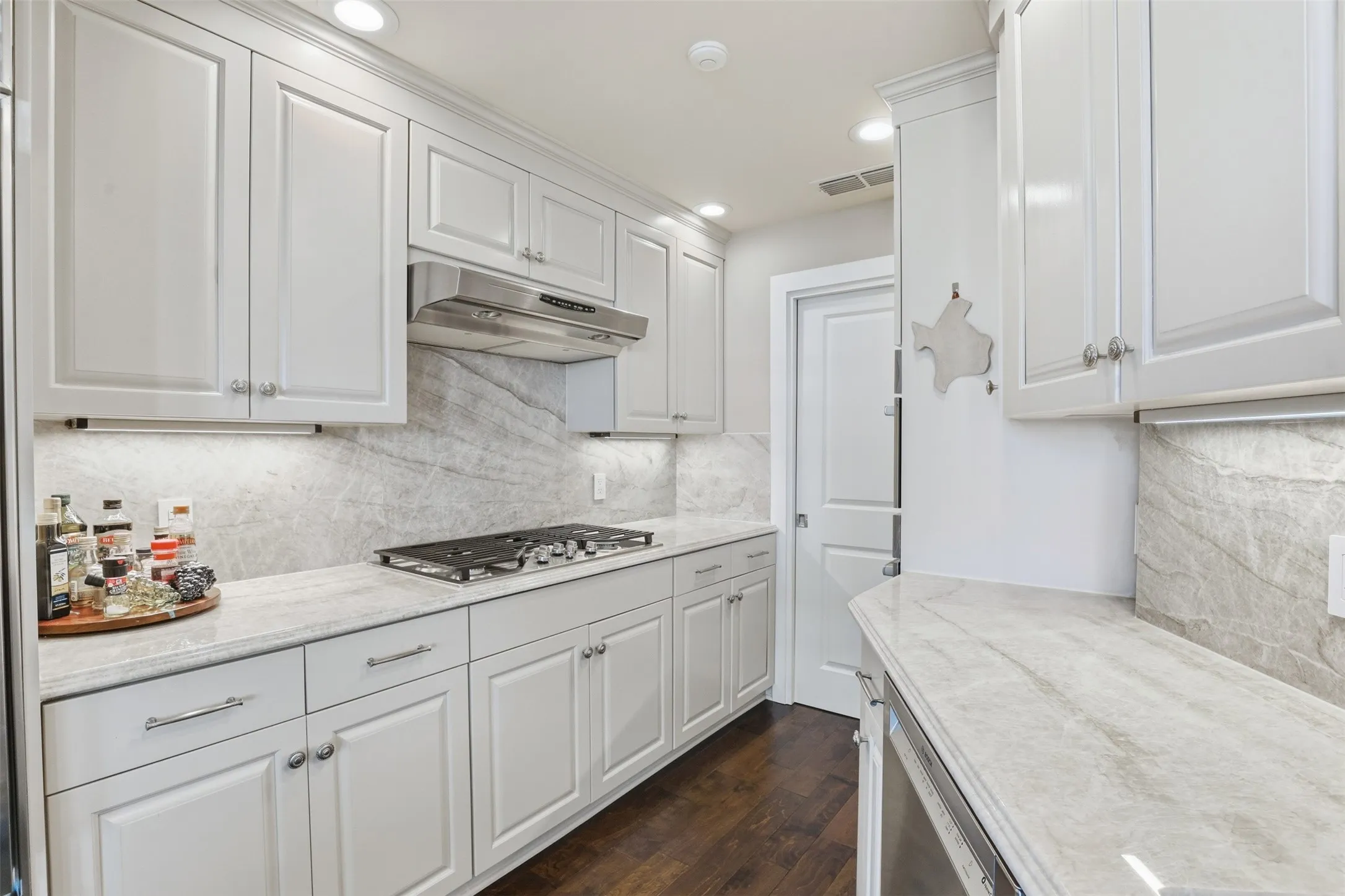 White cabinetry with polished hardware, stainless steel range hood, built-in cooktop, stone-finish countertops, and wood-finish flooring