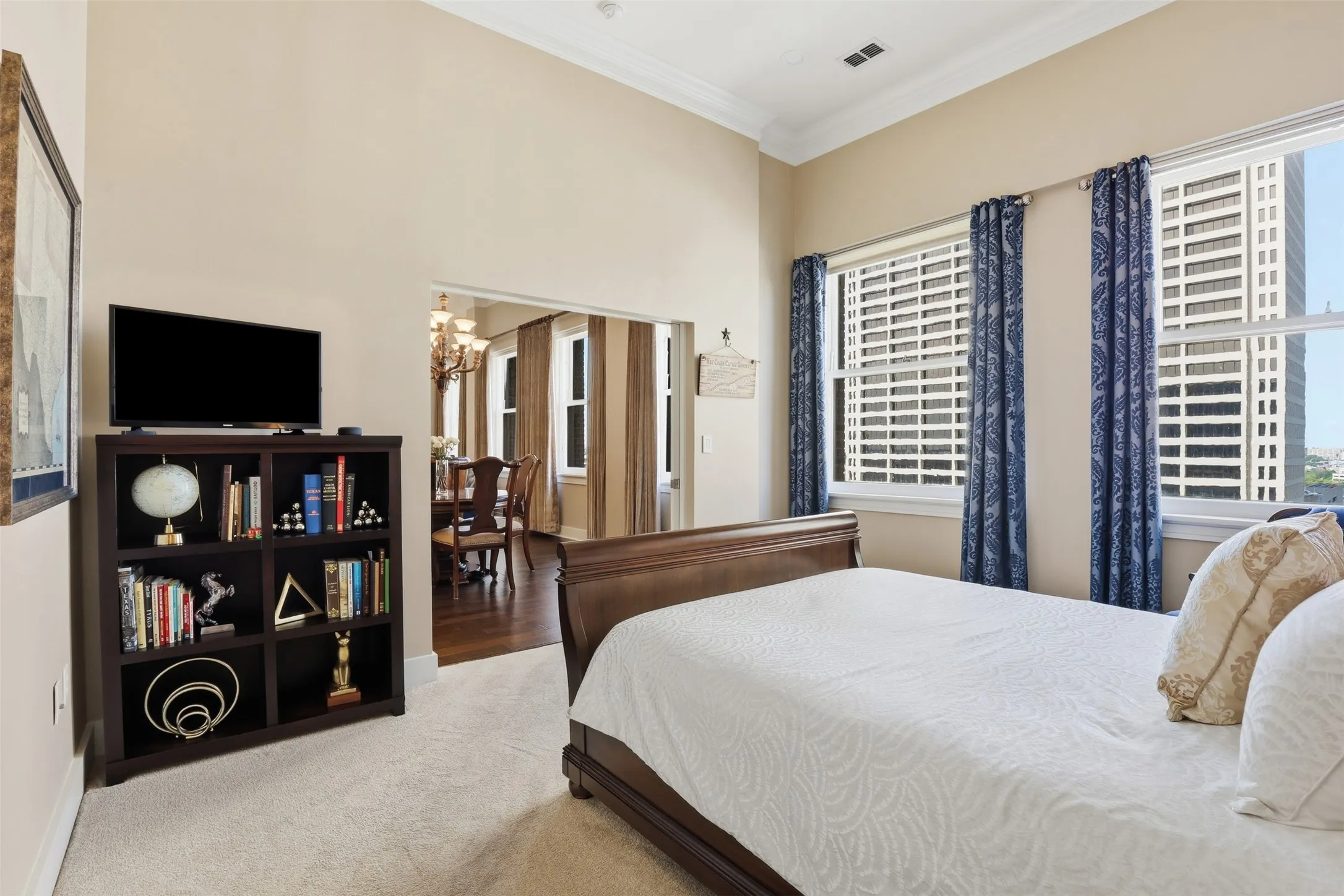 High ceilings with crown molding, light-toned wall paint, large windows with white blinds, and an open doorway reveal wood-finish flooring in an adjoining room