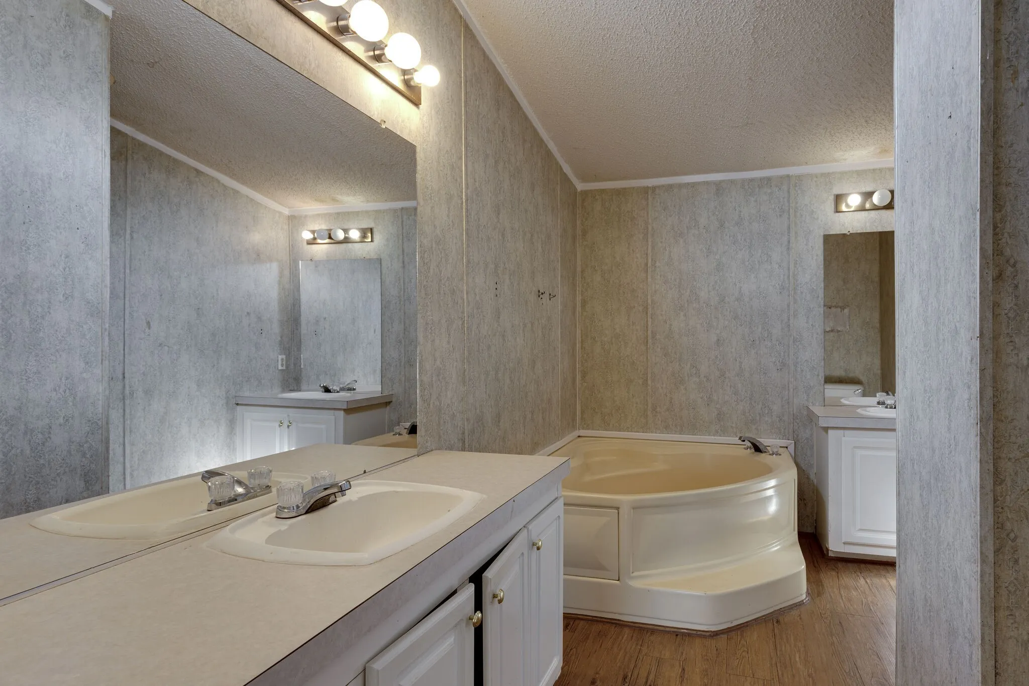 Dual vanity sinks with chrome fixtures, large wall-mounted mirror, built-in corner soaking tub, wood-finish flooring, and wall-mounted light fixtures