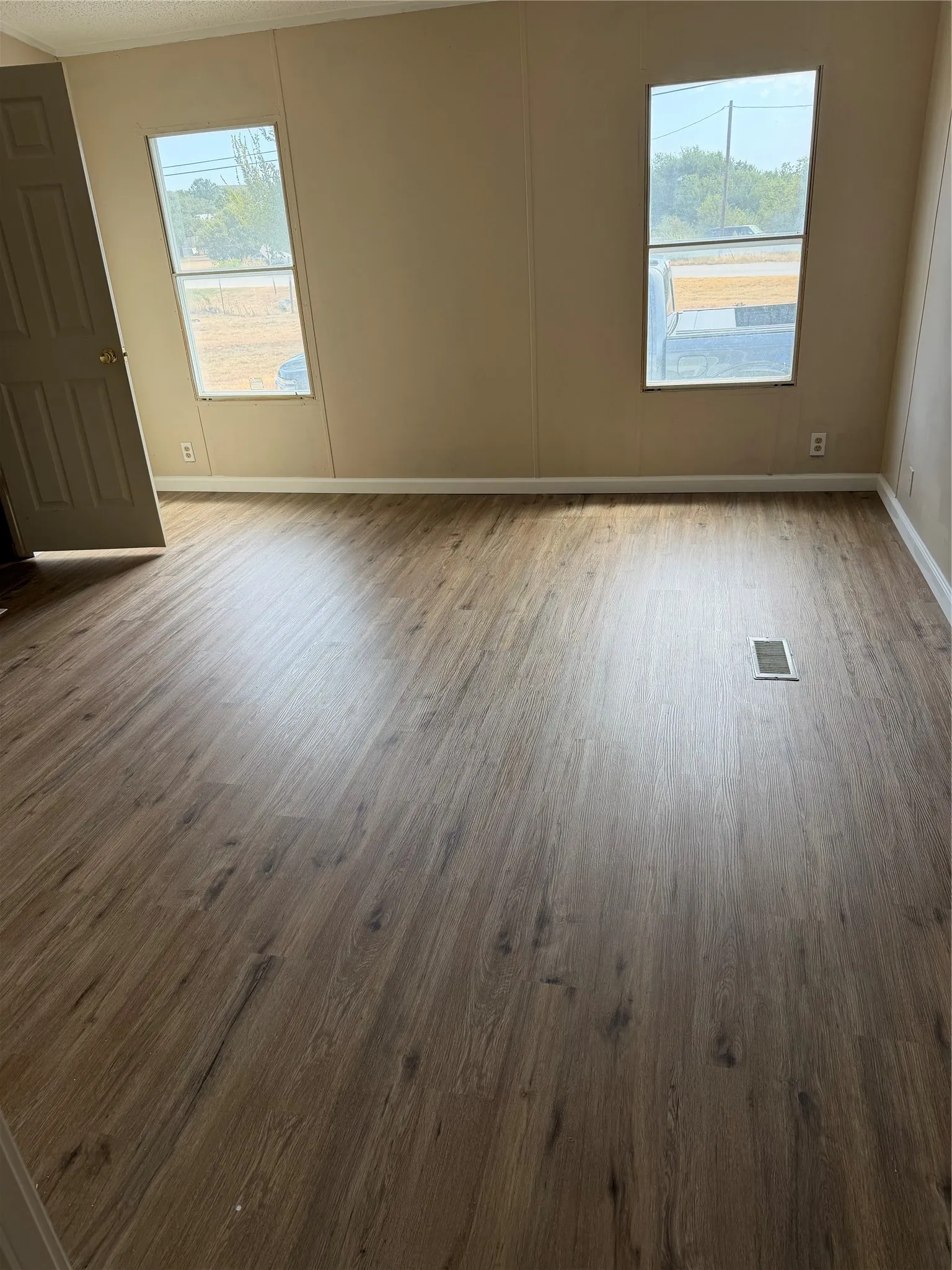 Spacious room flooded with natural light through dual windows, featuring wood-finish plank flooring and neutral wall finishes