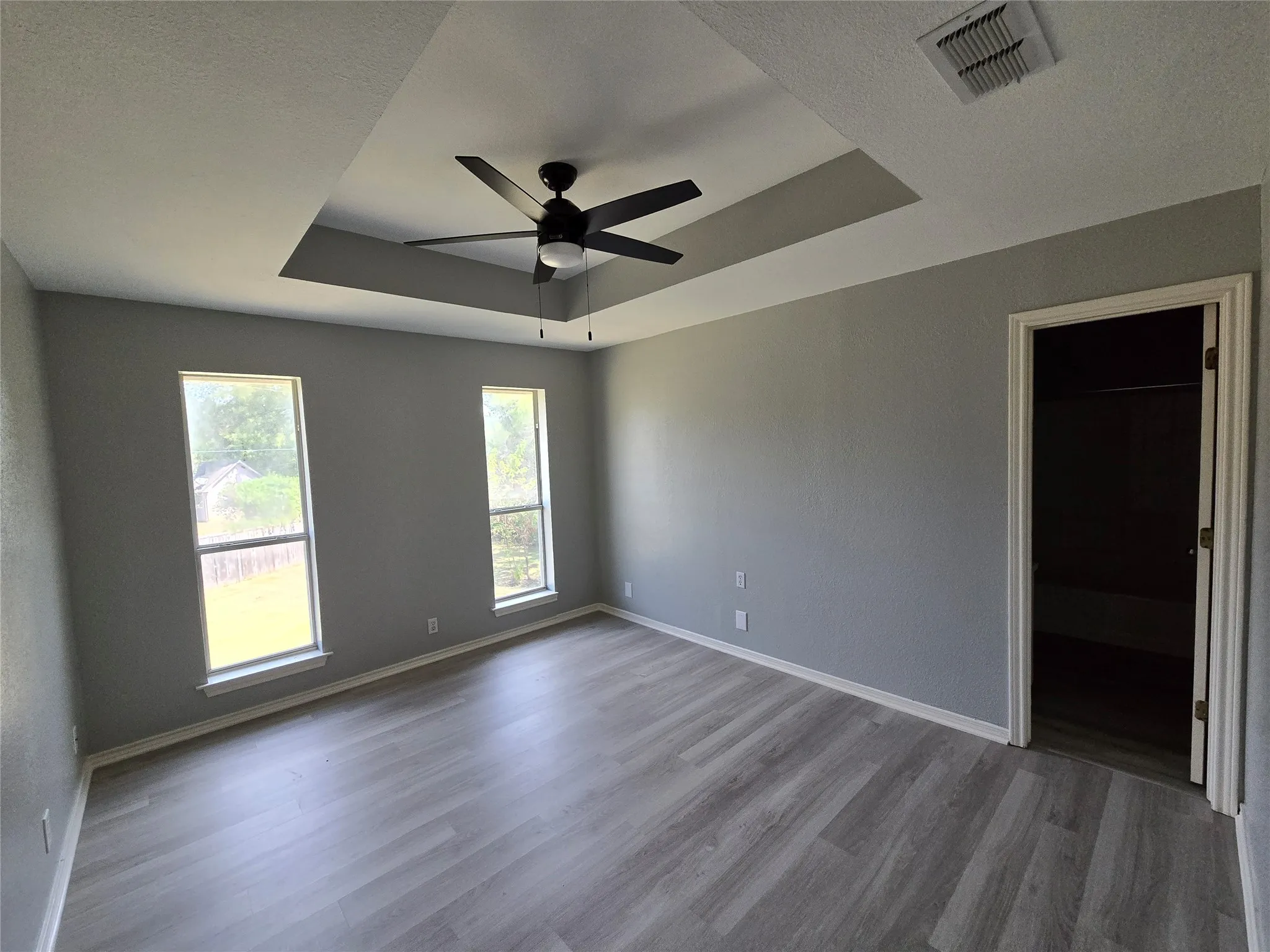 Tray ceiling with a black ceiling fan, two windows, gray walls, and wood-finish flooring