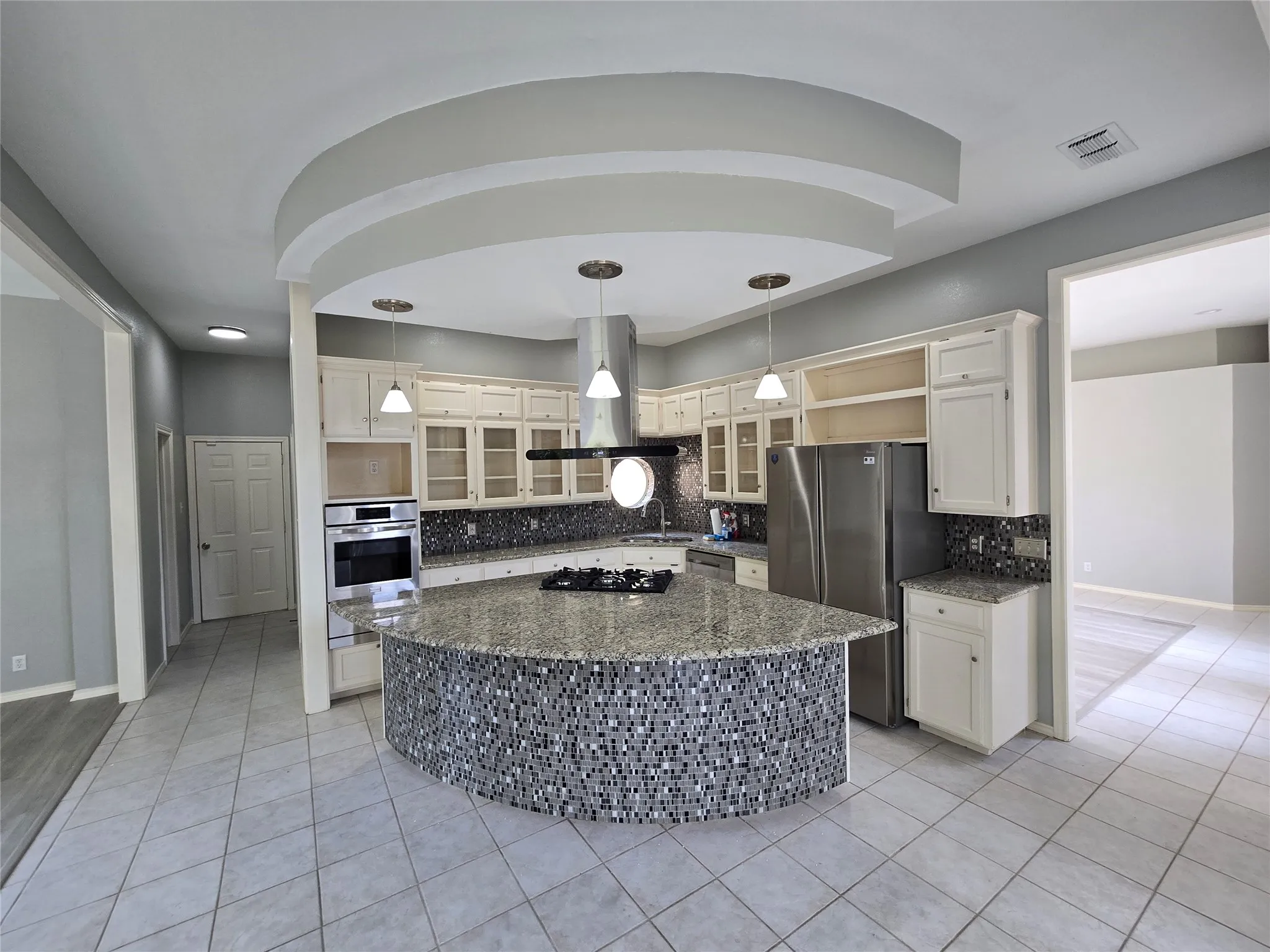 Gourmet kitchen featuring a curved granite-topped island with seating, stainless steel appliances, white cabinetry with glass-front upper storage, black subway tile backsplash, and pendant lighting