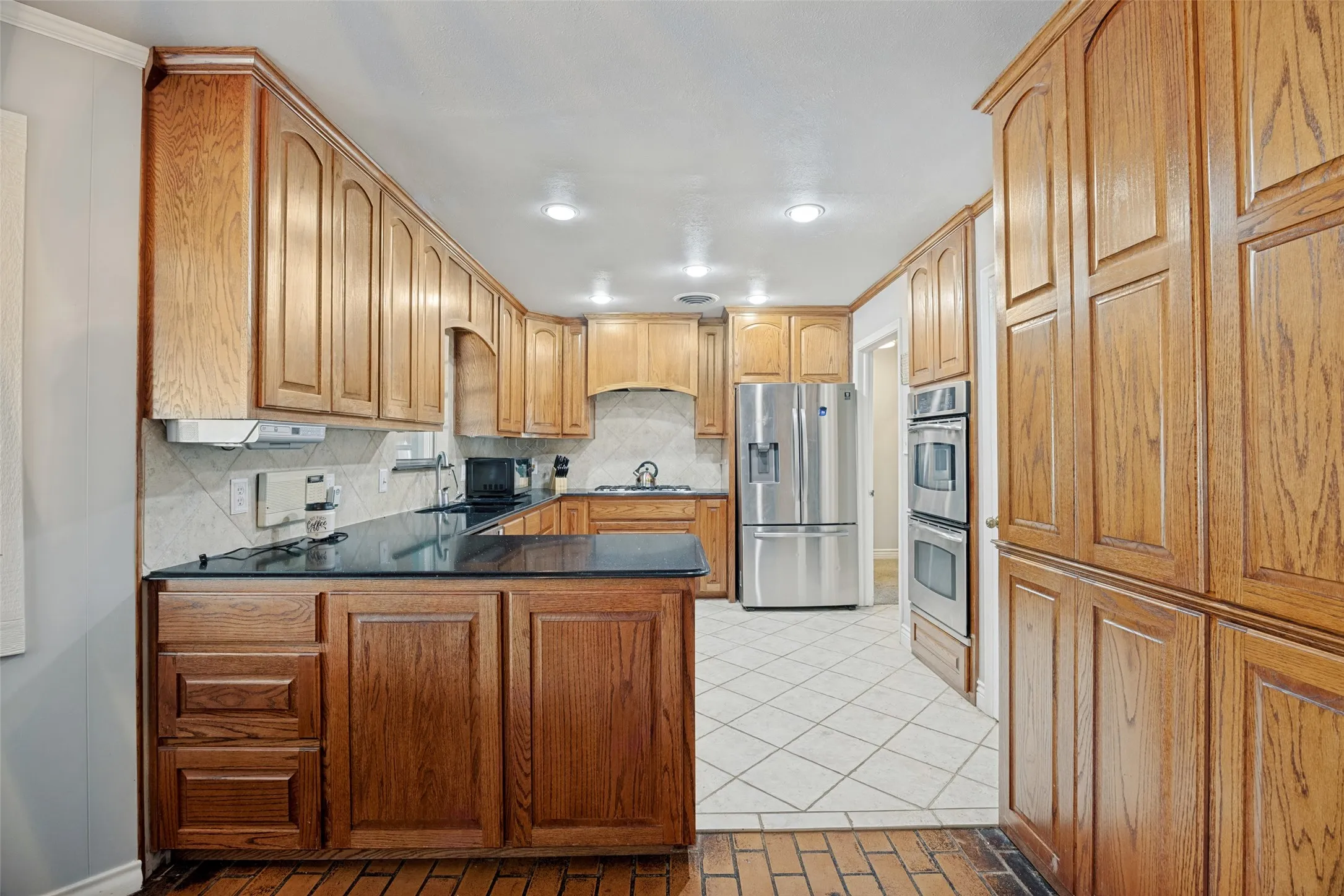 Kitchen featuring wood-finish cabinetry, dark countertops, a tiled backsplash, and stainless steel appliances including a refrigerator and double ovens