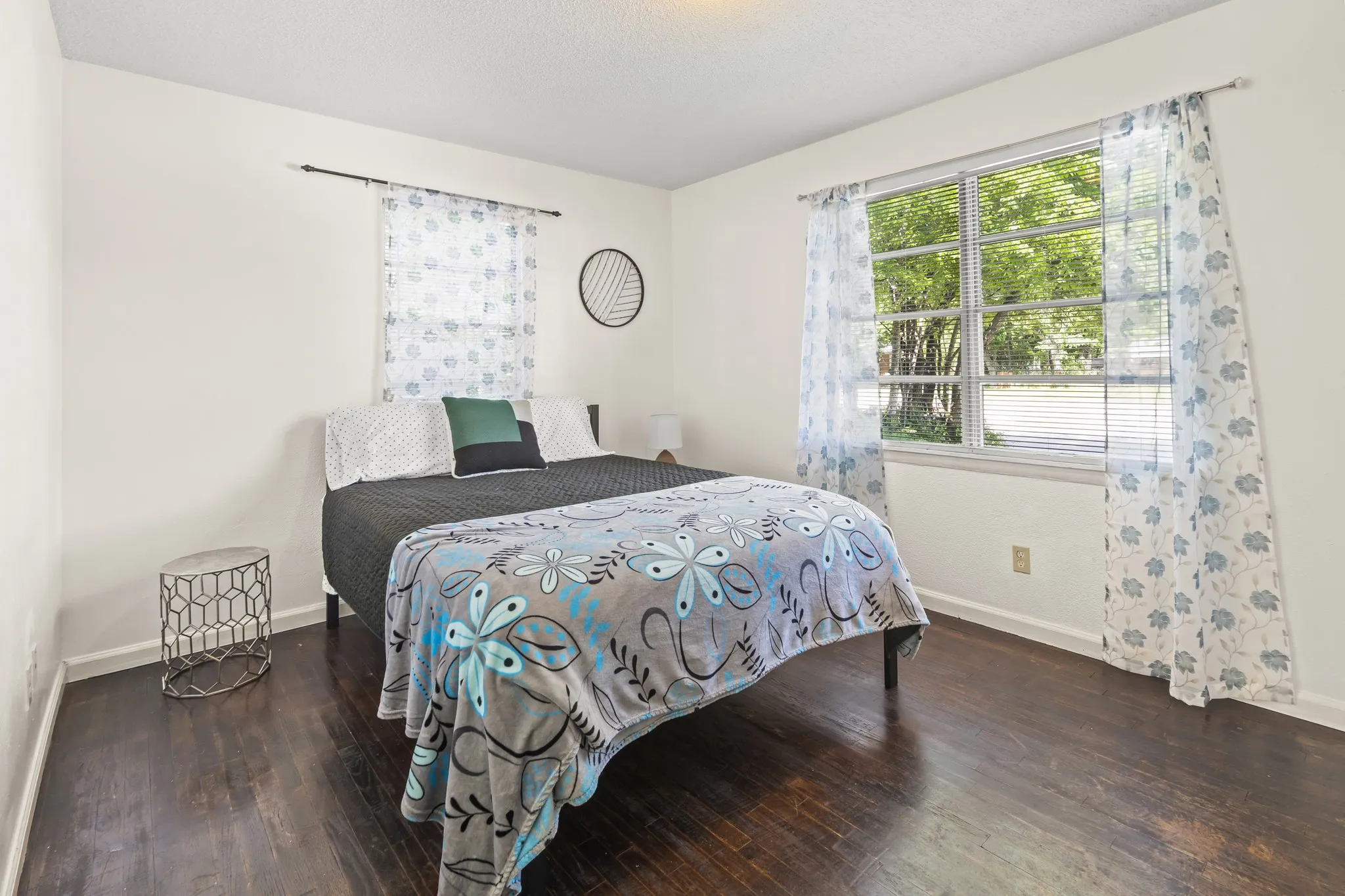 Bright secondary bedroom featuring hardwood flooring, large windows with natural light, sheer patterned curtains, and a decorative wall clock
