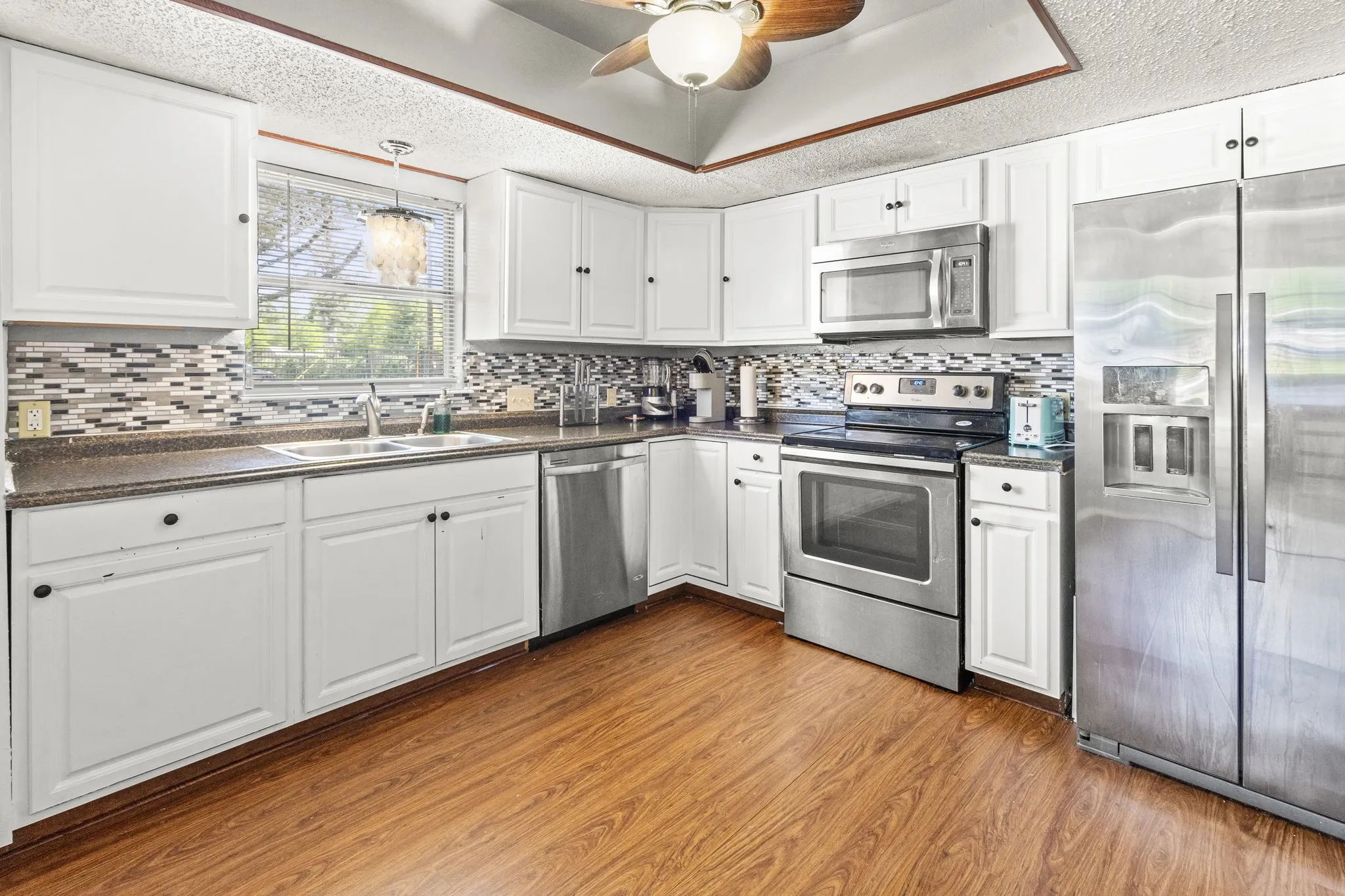 White cabinetry, stainless steel appliances, wood-finish flooring, mosaic tile look backsplash