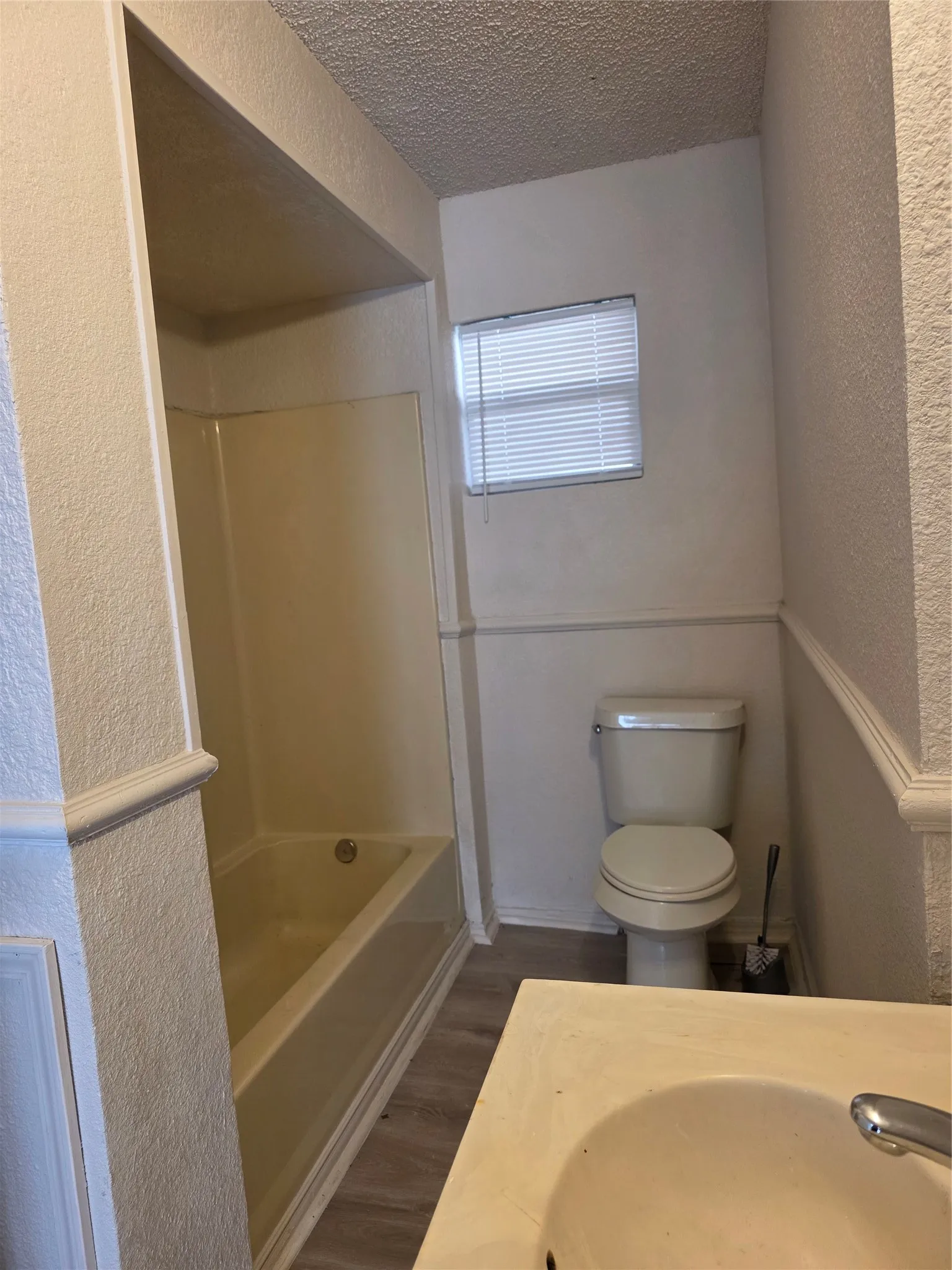 Tub/shower combination with integrated surround, wall-mounted sink, toilet, window with blinds, and wood-finish flooring