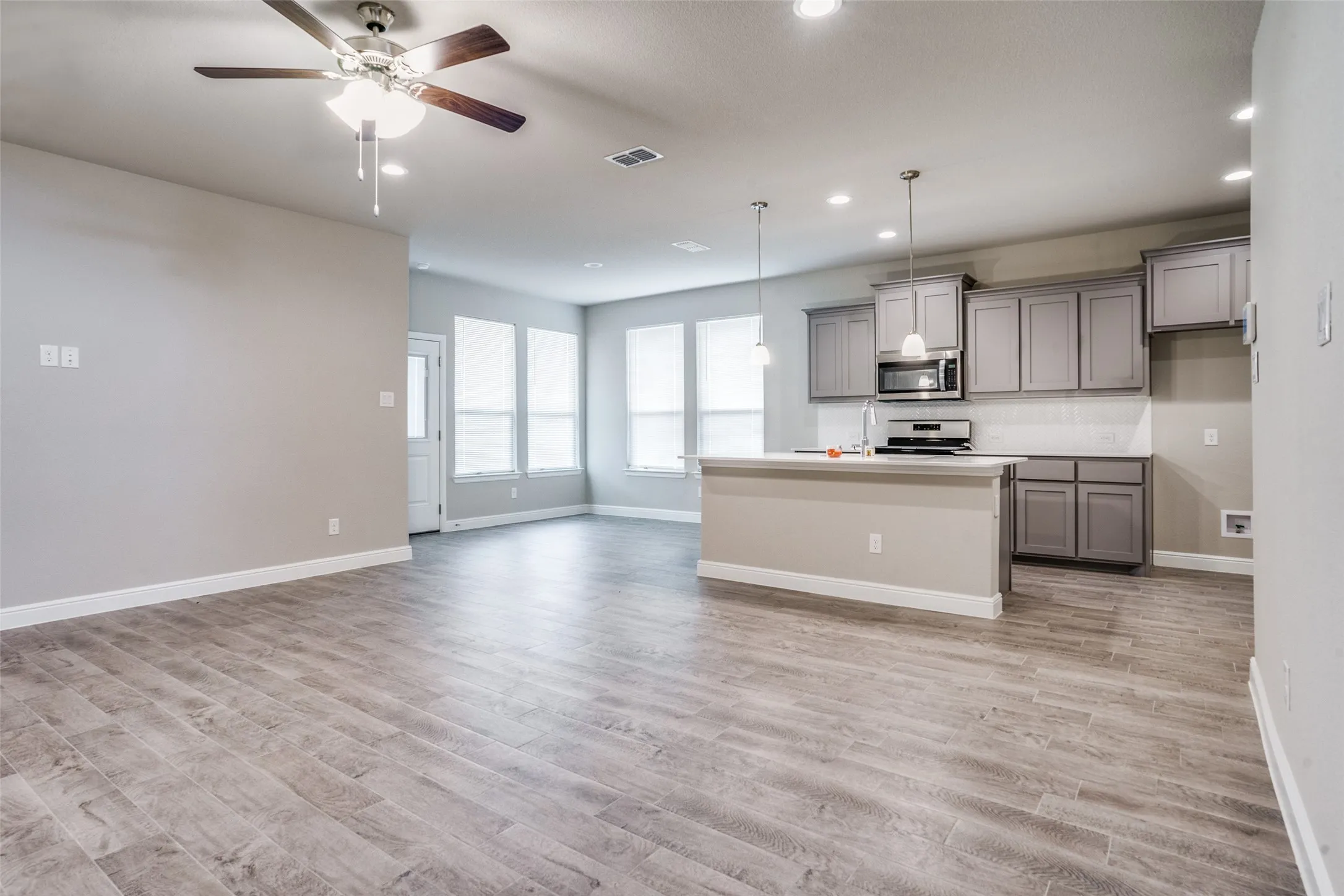 Wood-finish flooring throughout, kitchen with gray cabinetry, white countertops, and stainless steel appliances, and multiple windows with blinds